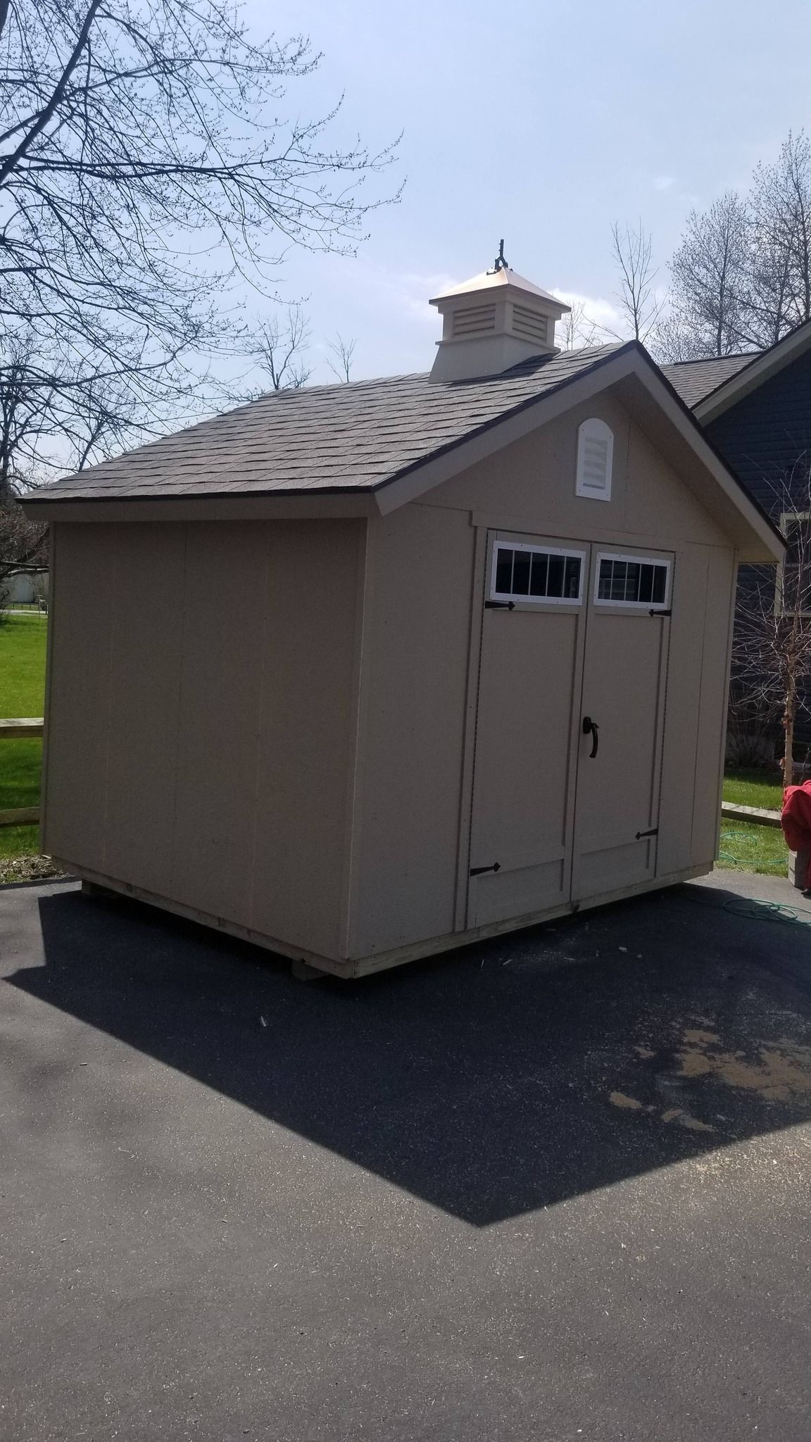 Small beige garden shed with double doors and windows on a driveway, surrounded by trees and blue sky