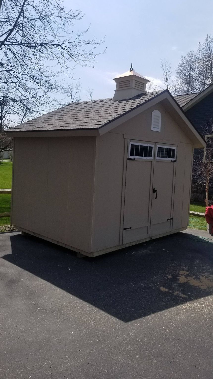 Small beige garden shed with double doors and windows on a driveway, surrounded by trees and blue sky