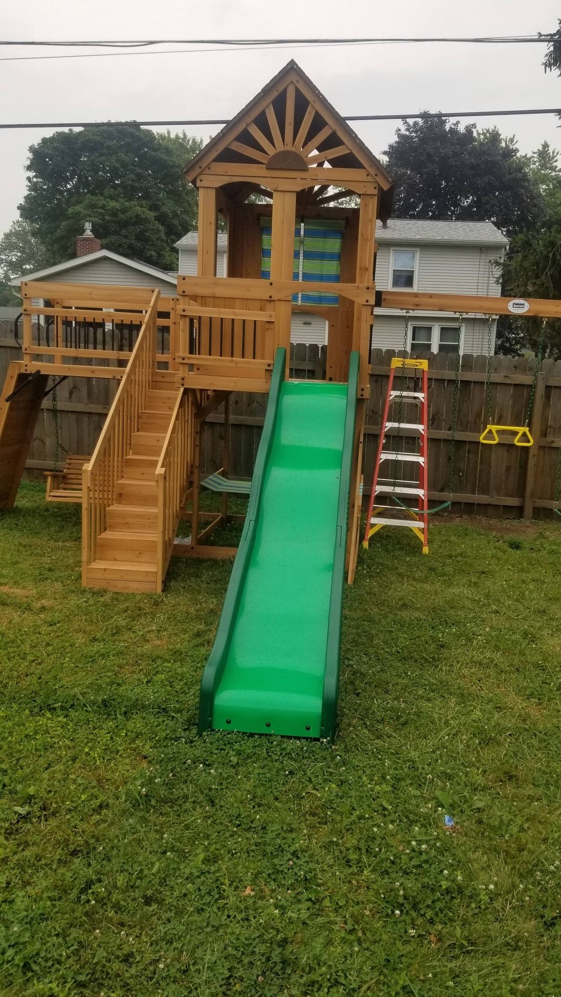 Wooden backyard playset with green slide, stairs, and ladder on a grassy lawn