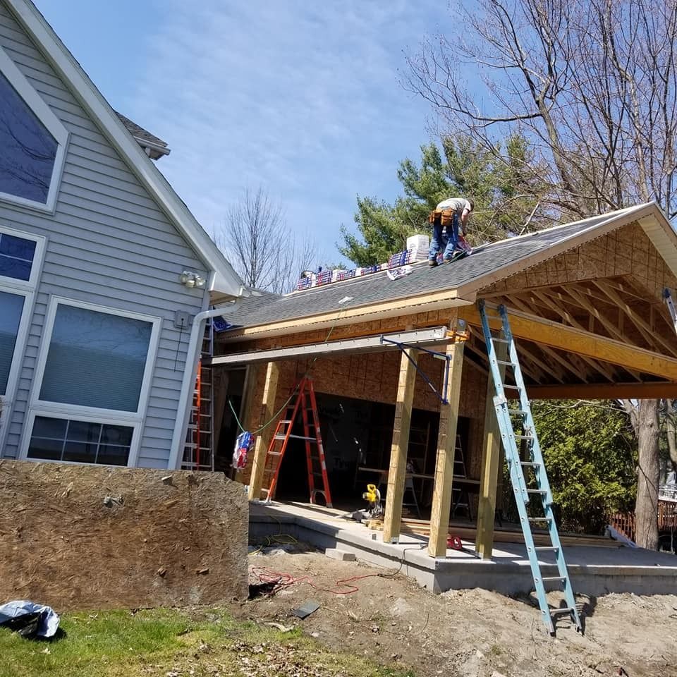 Workers installing shingles on a new carport roof beside a house, with ladders and exposed beams.