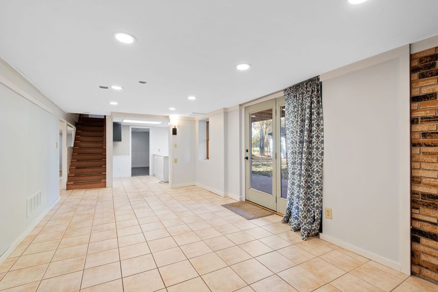 Bright tiled empty room with white walls, recessed lights, stairs, and sliding glass doors with curtains