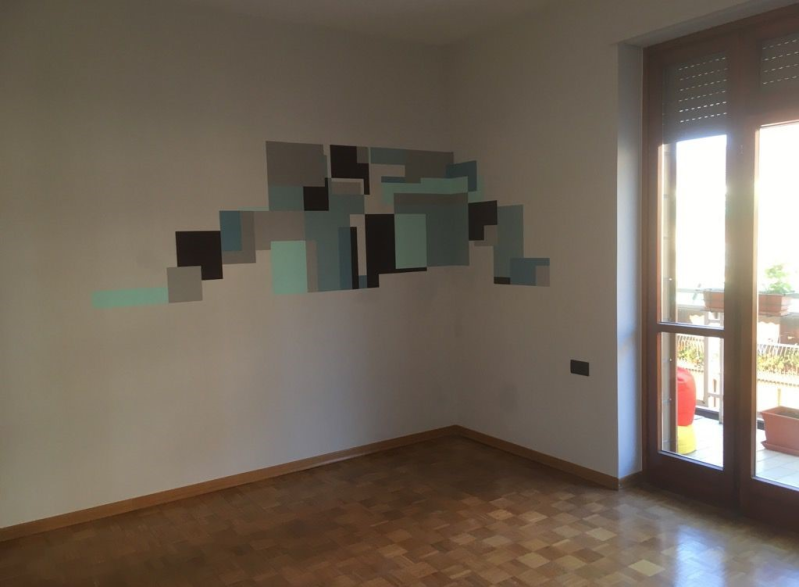 Arte murale astratta raffigurante quadrati dipinti in varie tonalità di grigio e blu, in una stanza vuota con una finestra.