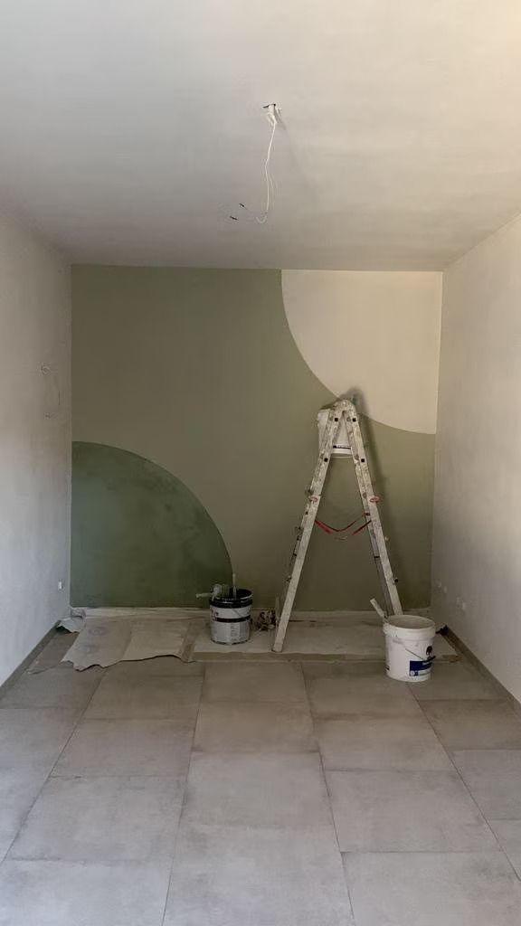 Stanza con parete dipinta di verde e bianco, scala, barattoli di vernice e pavimento piastrellato; stanza in fase di ristrutturazione.