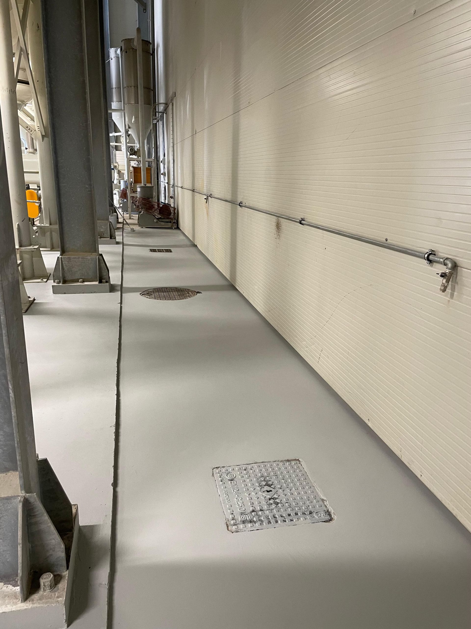 Lungo corridoio industriale grigio con tubi metallici lungo le pareti e grate nel pavimento.