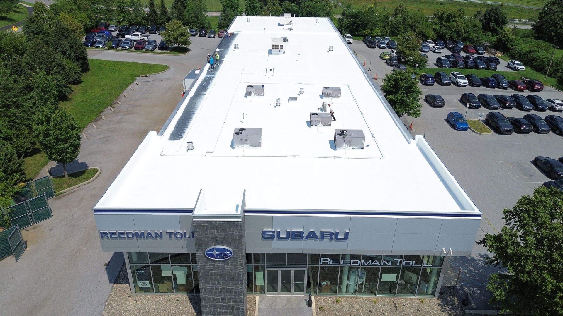 Aerial view of a white-roofed industrial building with several parked vans and loading docks in an asphalt lot.