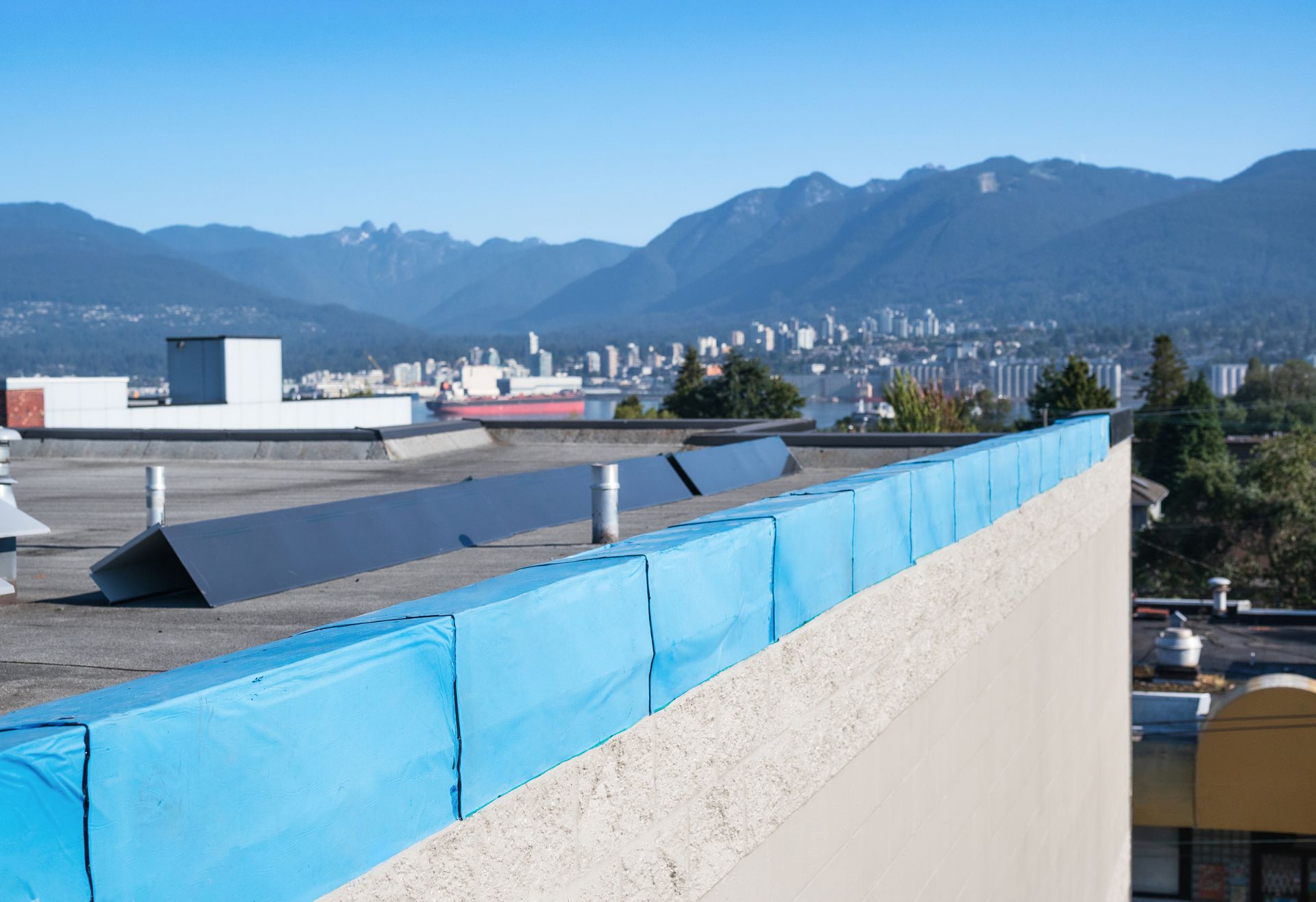 A flat building roof with a blue protective barrier along the edge, overlooking a city skyline and mountains.
