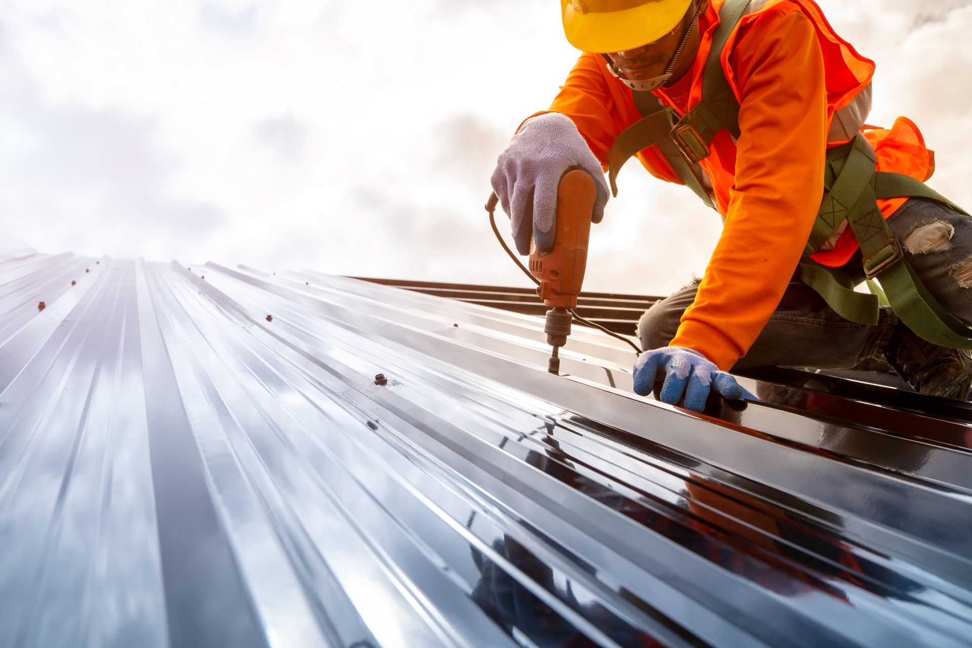 A construction worker in a safety harness and yellow hard hat uses a drill to secure metal roofing panels.