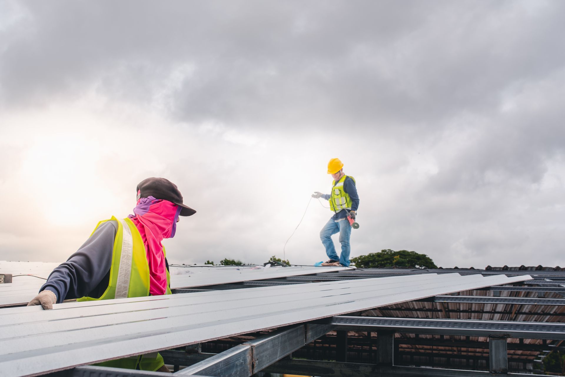 Two construction workers in safety vests and gear install metal roofing panels on an outdoor frame under a cloudy sky.