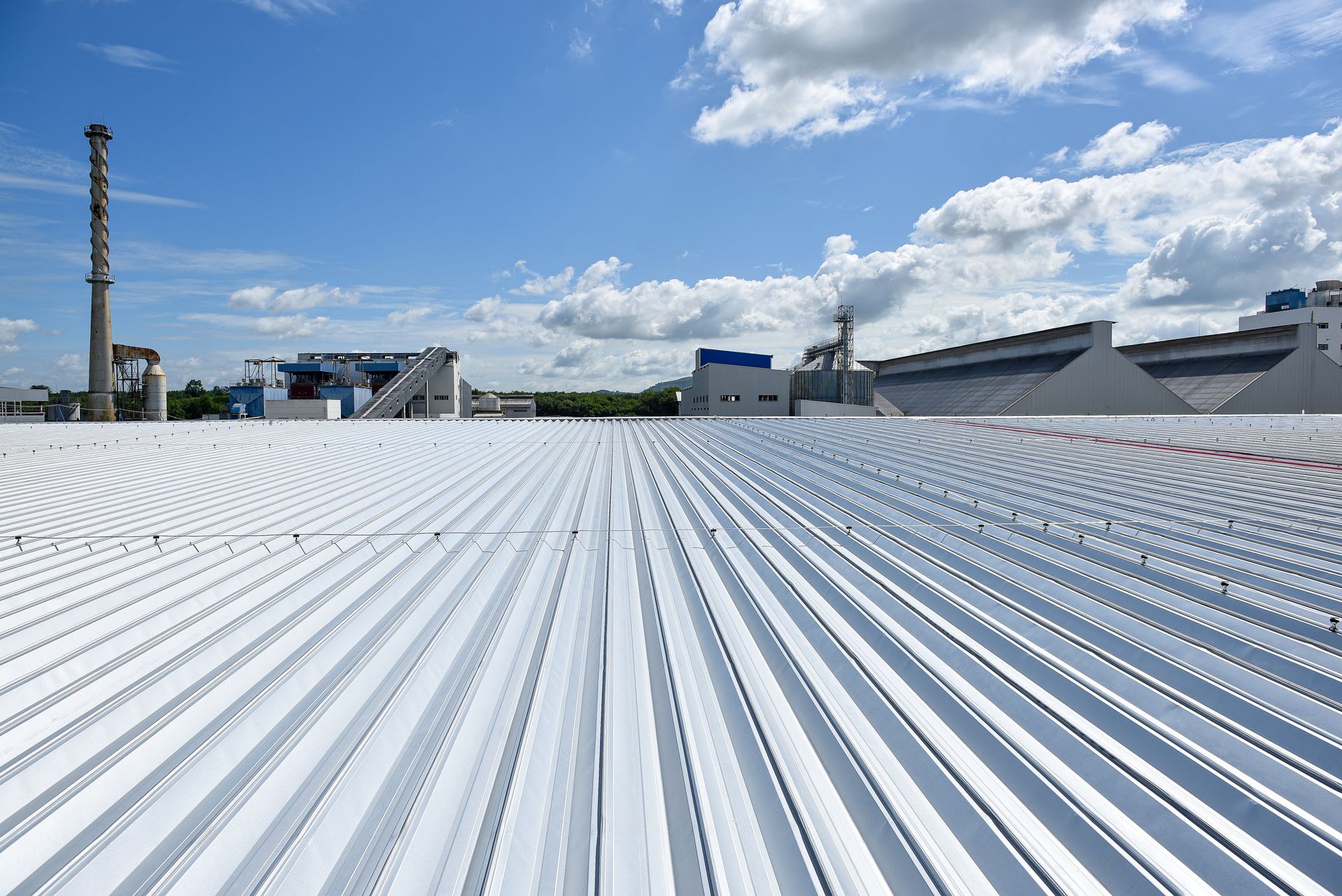A view across a large, white, corrugated metal industrial roof toward distant factory buildings under a blue sky.
