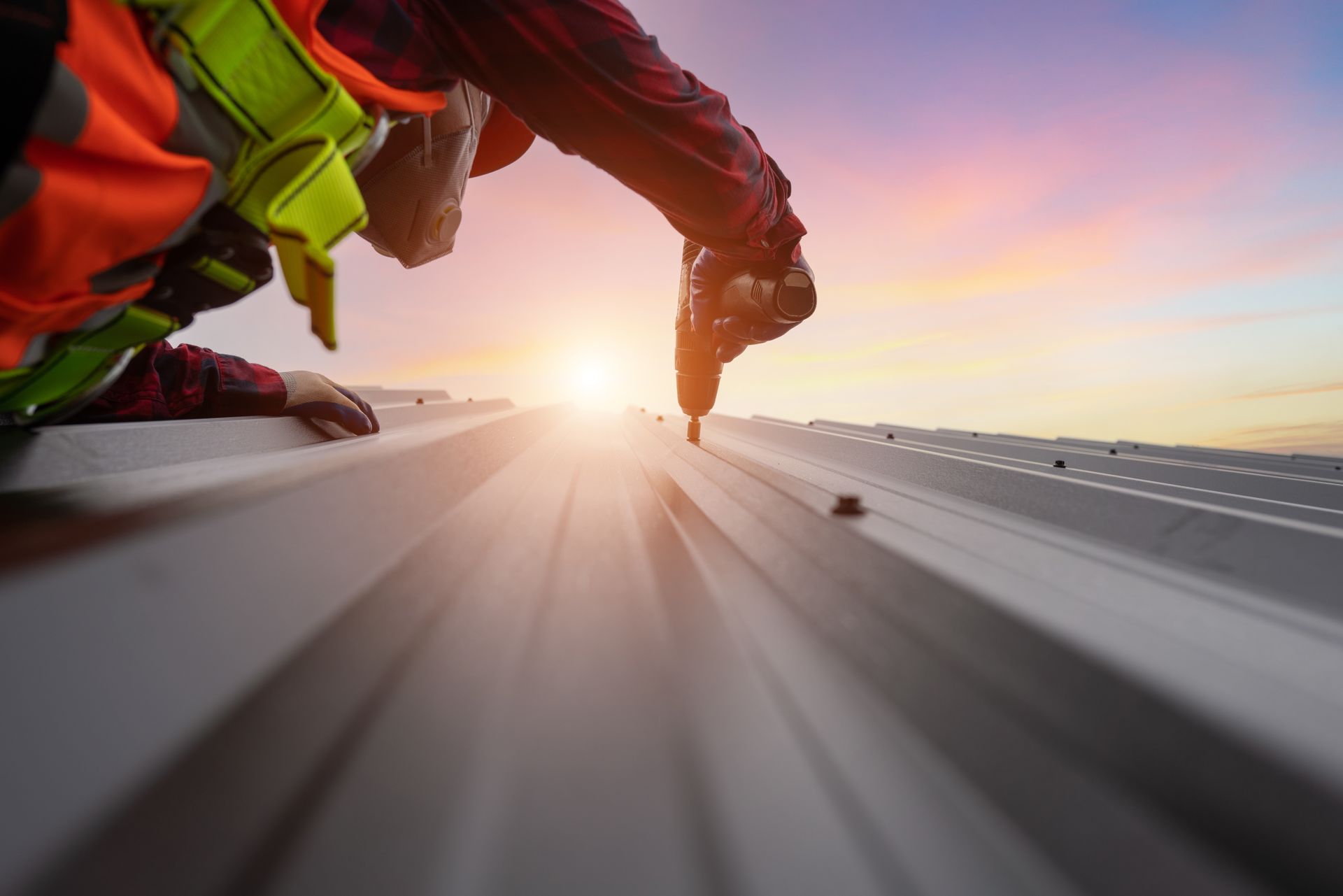 A worker in high-visibility gear uses a power drill to fasten metal panels on a roof at sunset.