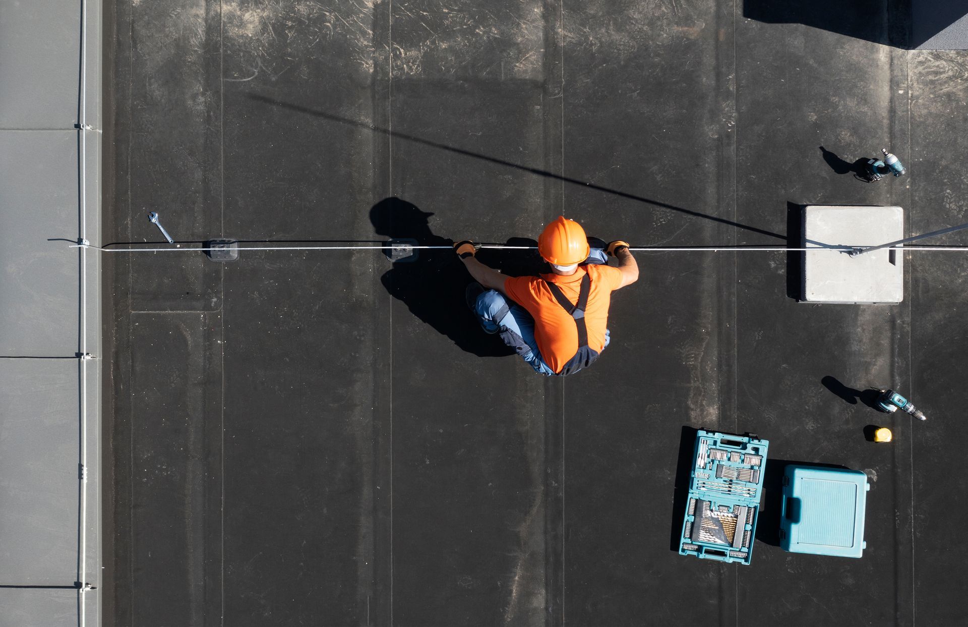 An aerial view of a worker in an orange shirt and harness installing a cable across a black flat roof.