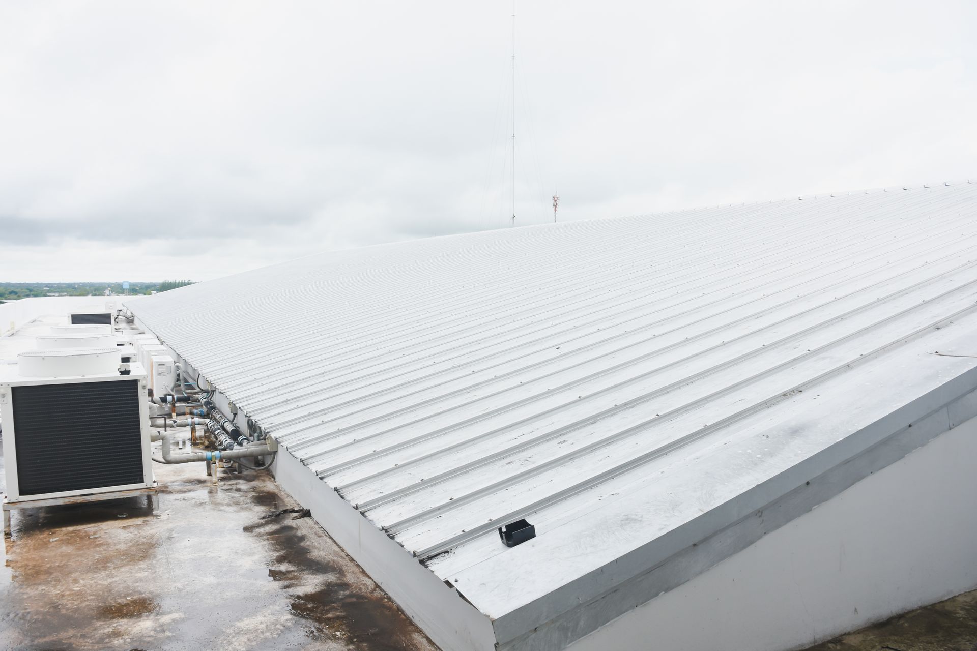 A white corrugated metal roof on a building, with an HVAC unit on the left and a gray overcast sky above.