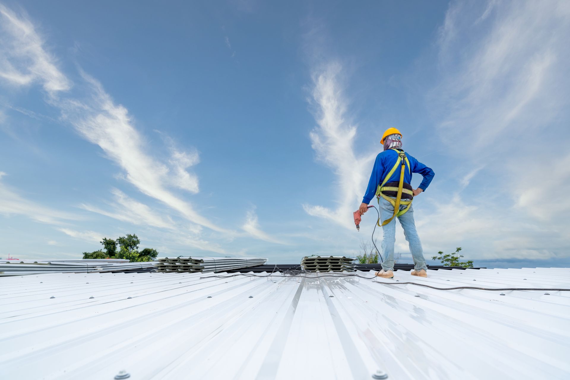 A construction worker in a yellow hard hat and safety harness stands on a white industrial metal roof under a blue sky.
