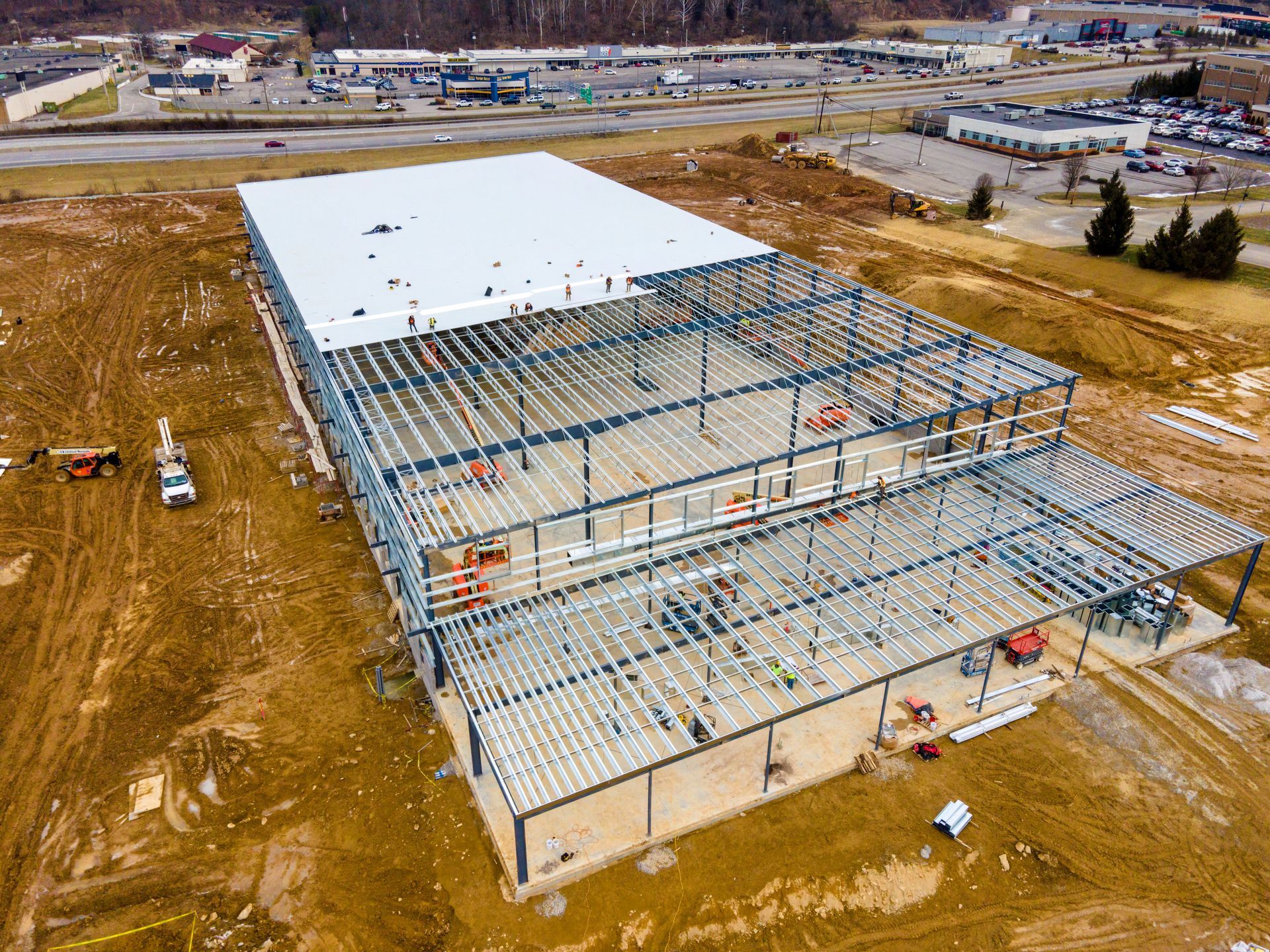 Aerial view of a large commercial building under construction with a steel frame and partially installed white roof.