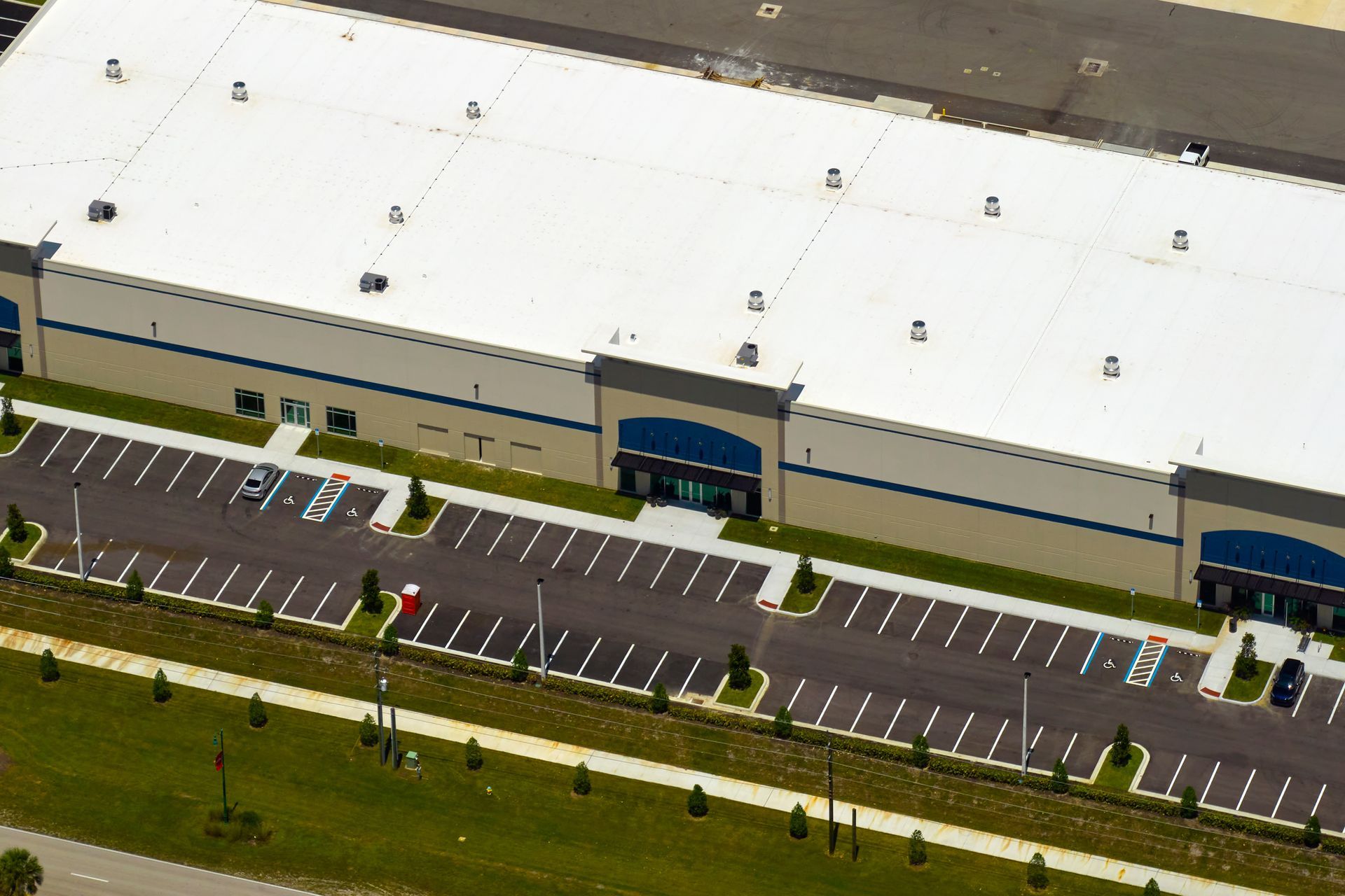 Aerial view of a large, beige industrial warehouse with a white flat roof, blue accents, and an adjacent parking lot.