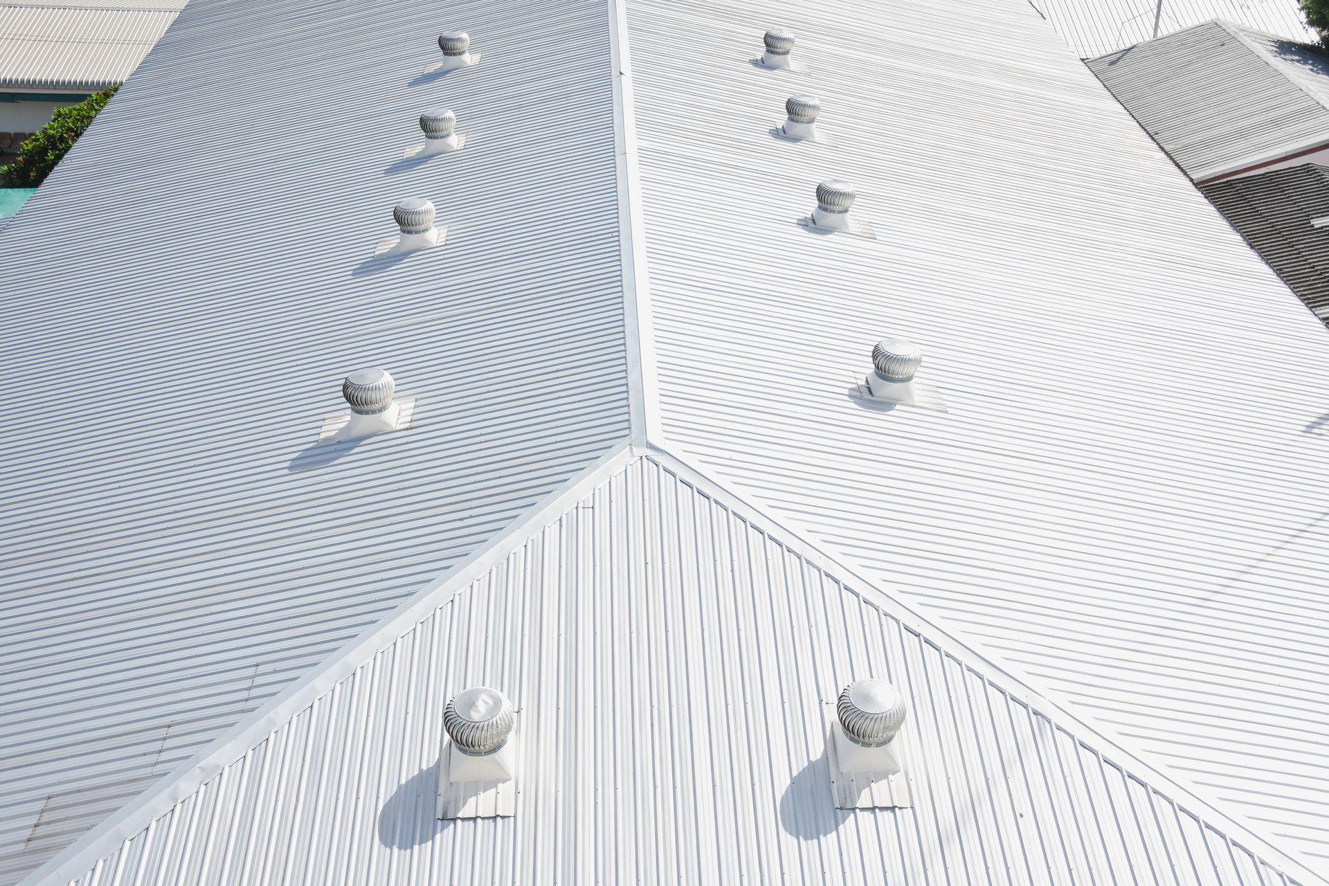 An aerial view of a white, corrugated metal roof with multiple spinning turbine roof vents installed on the slopes.