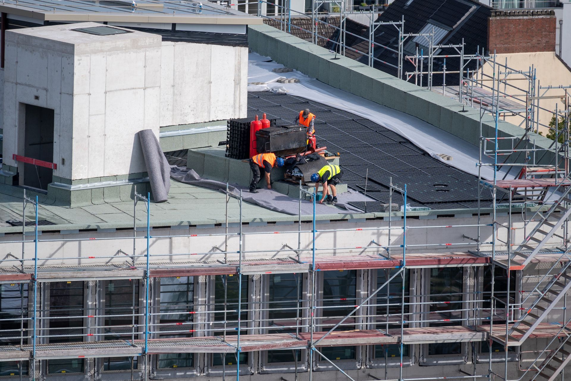 Workers in high-visibility vests install black roofing materials on a building roof surrounded by scaffolding.