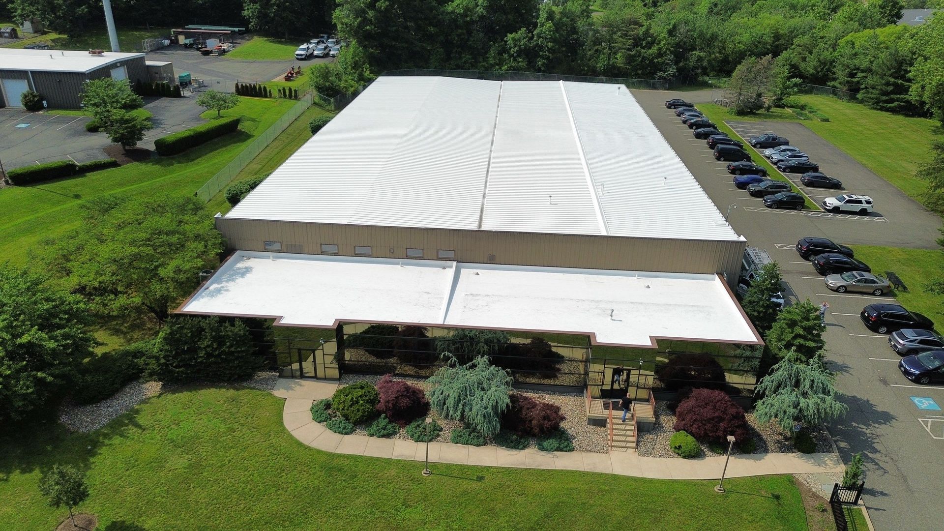 Aerial view of a white-roofed industrial building with several parked vans and loading docks in an asphalt lot.