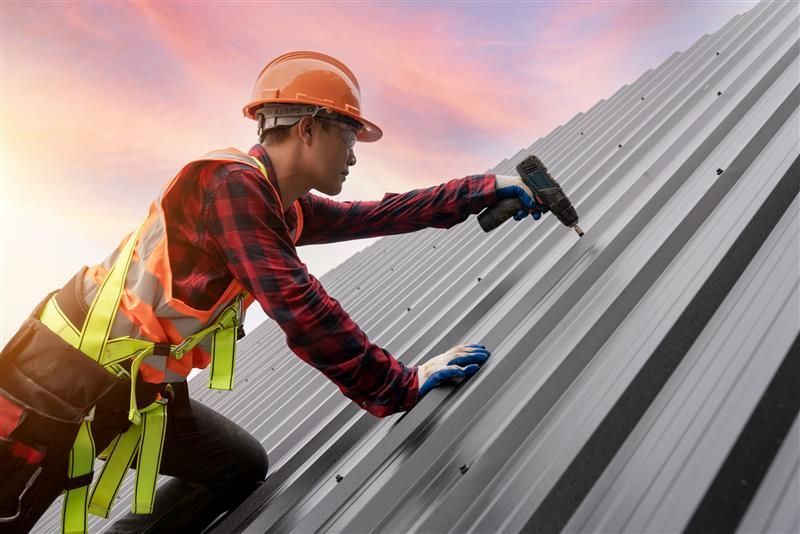 A worker in safety gear using a power drill on a metal roof during sunset.
