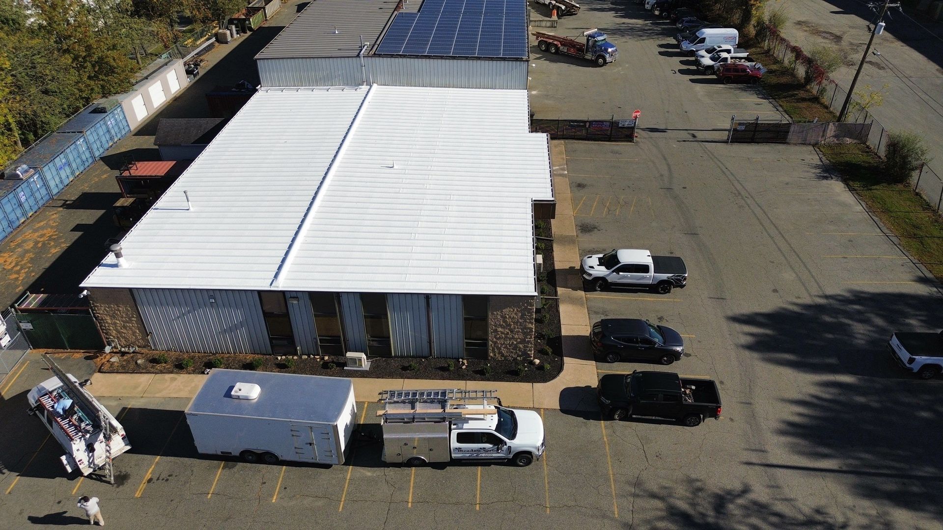 Aerial view of a white-roofed industrial building with several parked vans and loading docks in an asphalt lot.