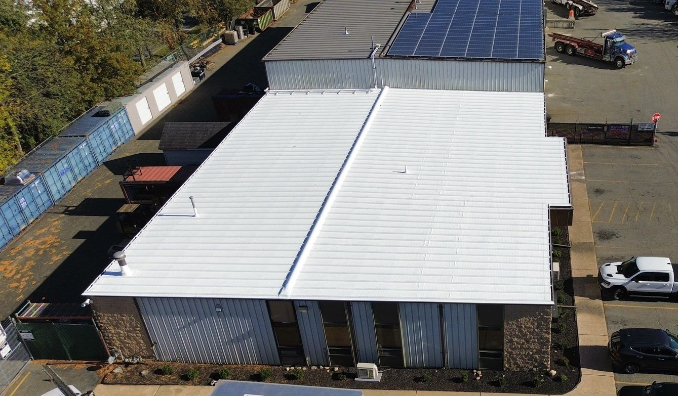 An aerial view of a building with a newly coated white metal roof, adjacent to a parking lot and solar panels.