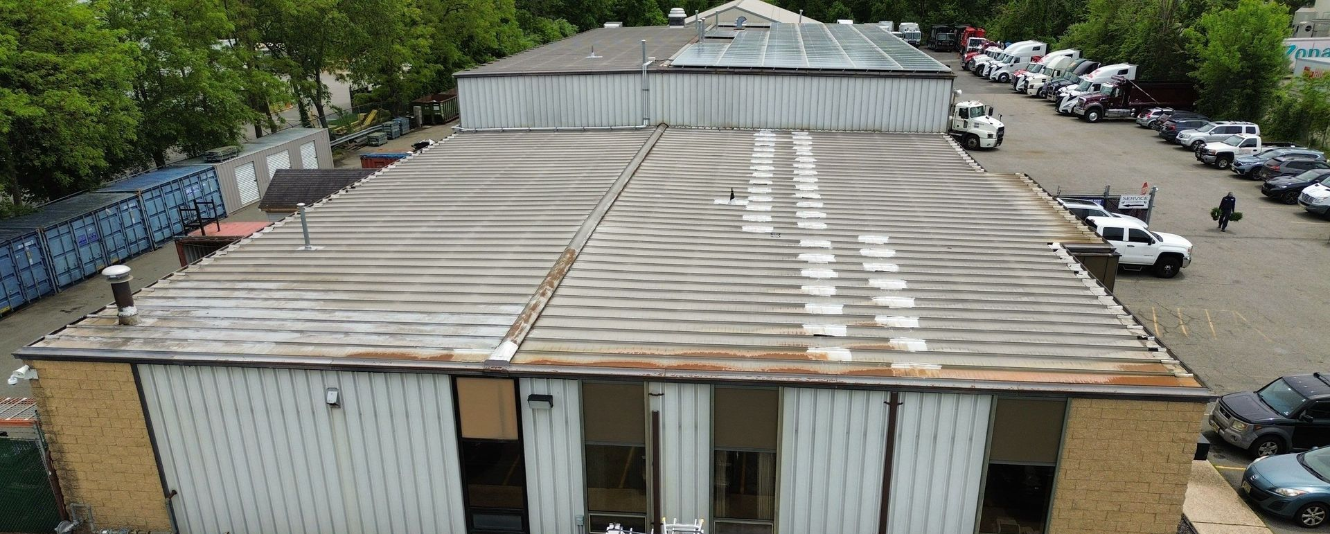 An aerial view shows a commercial building with a wavy, aged roof, adjacent to a parking lot filled with various cars.