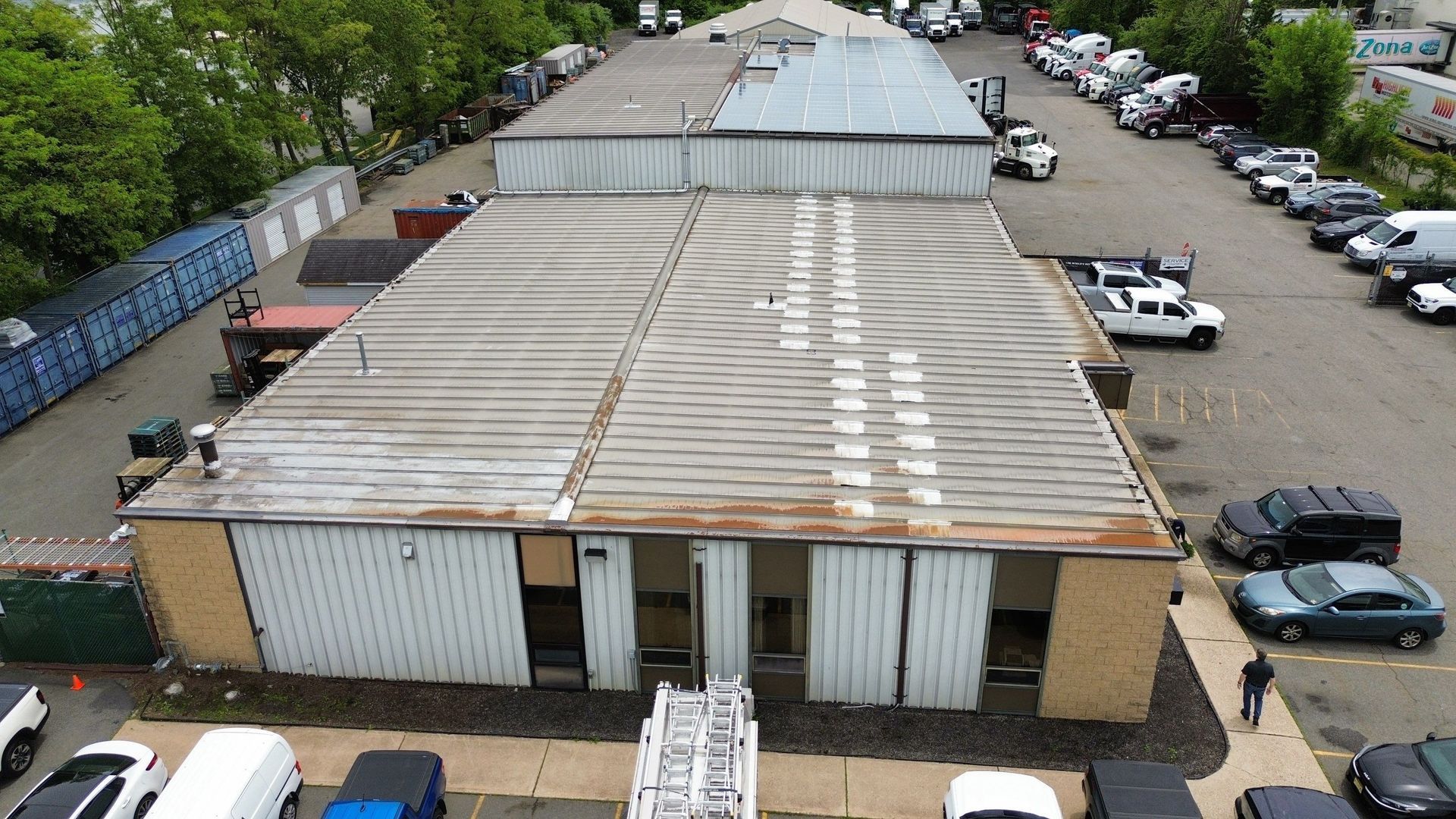 Overhead view of a gray warehouse with a curved metal roof, a yellow tanker truck, and green foliage nearby.