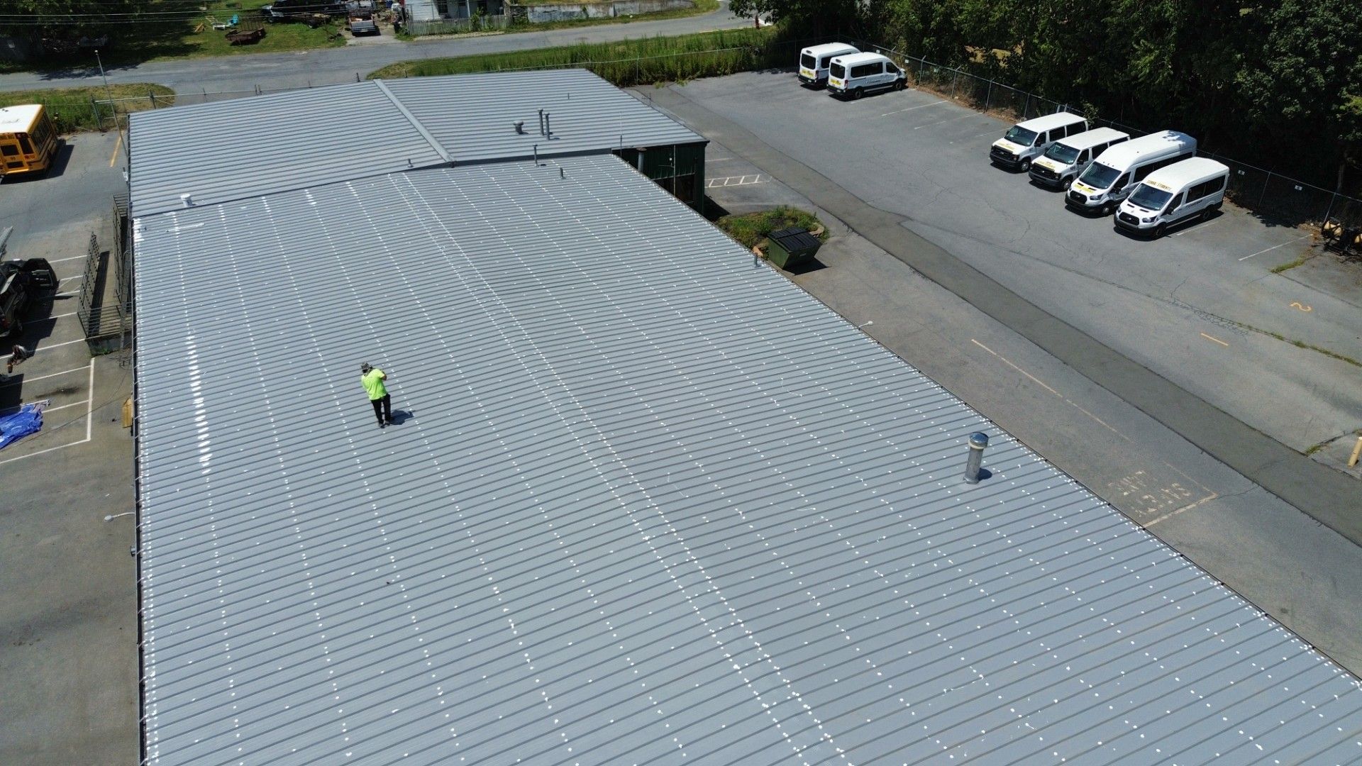 Workers on a large, curved metal roof, with grid pattern of solar panels, in front of parked vehicles on a sunny day.