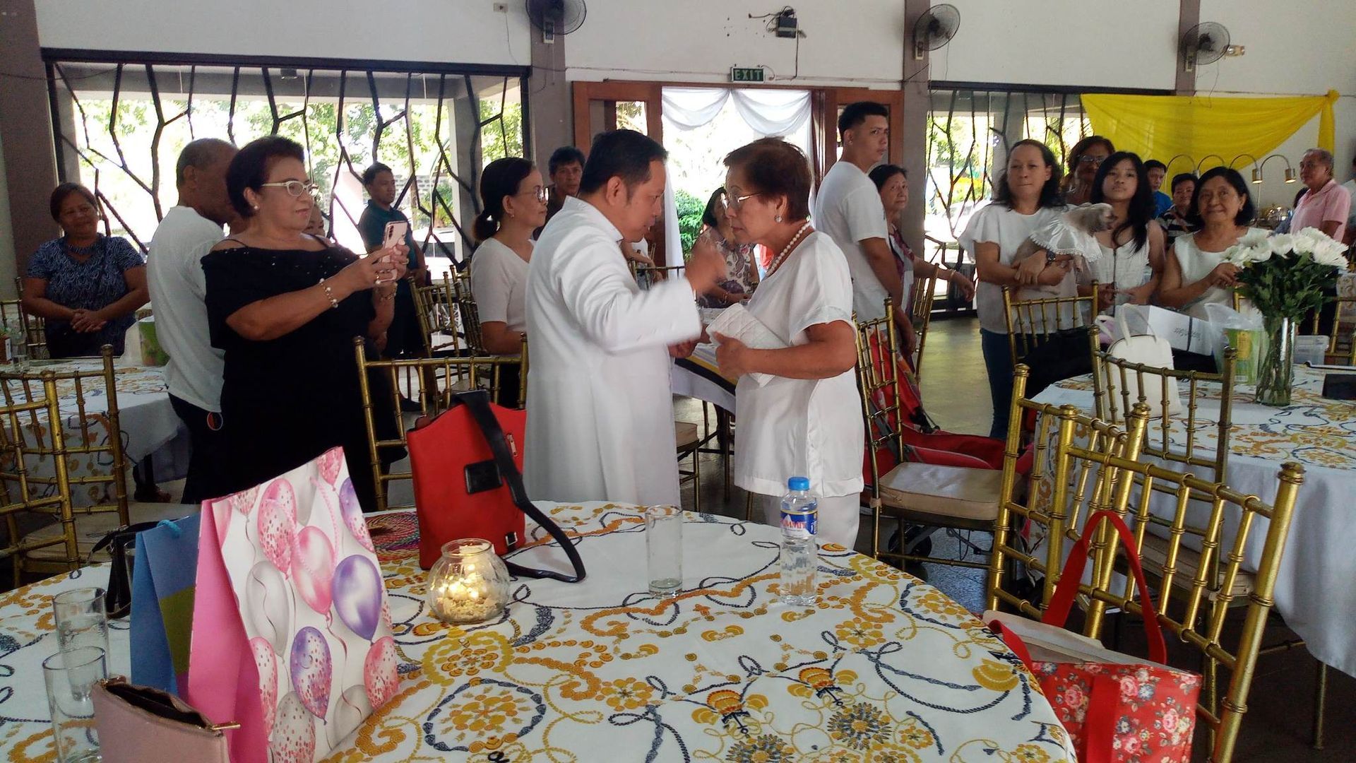 Group celebrates in a decorated room, a priest and woman embrace near a table, others watch.