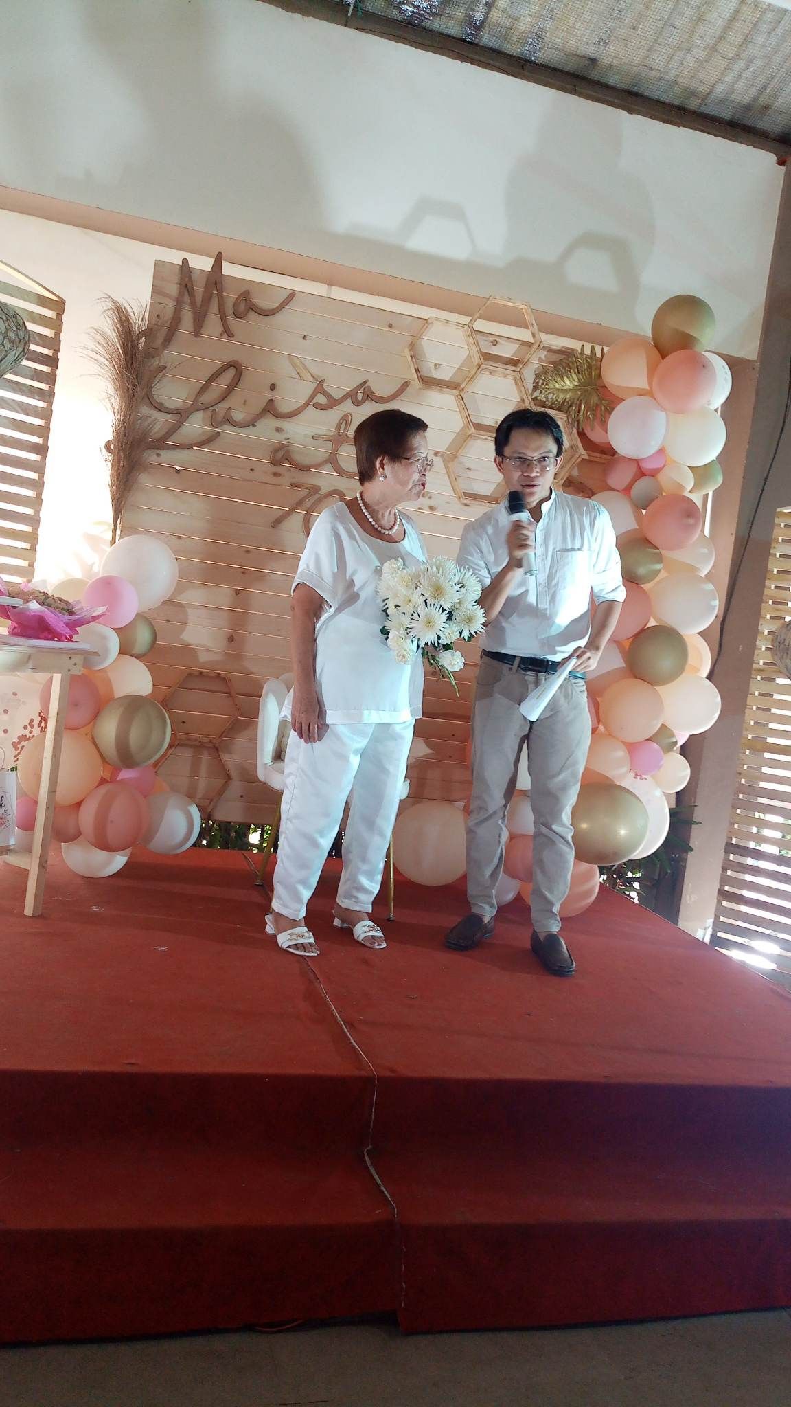 Two people on stage with balloons; woman in white, man with microphone speaking.
