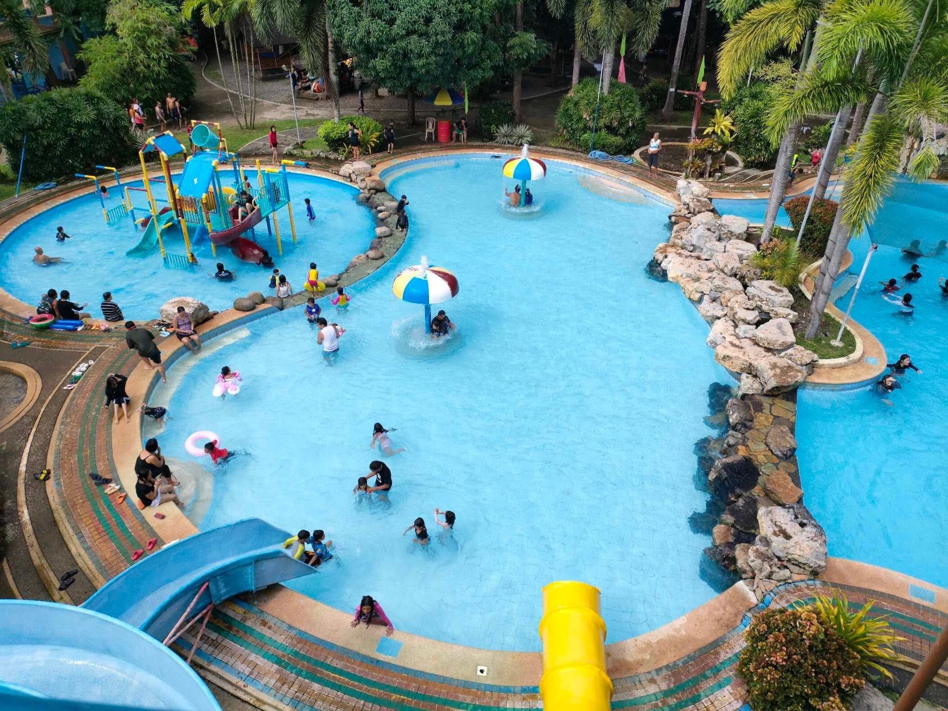 Water park with multiple pools, waterslides, and people swimming. Blue water, green trees, and a sunny day.