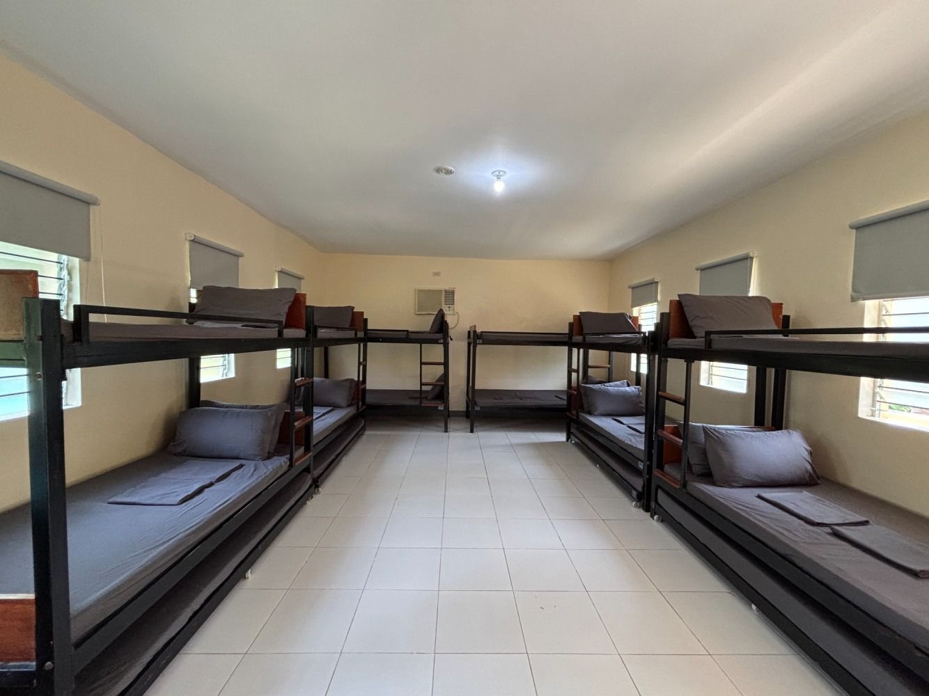 Dorm room with bunk beds; beige walls, grey mattresses, and window blinds, with a few windows.