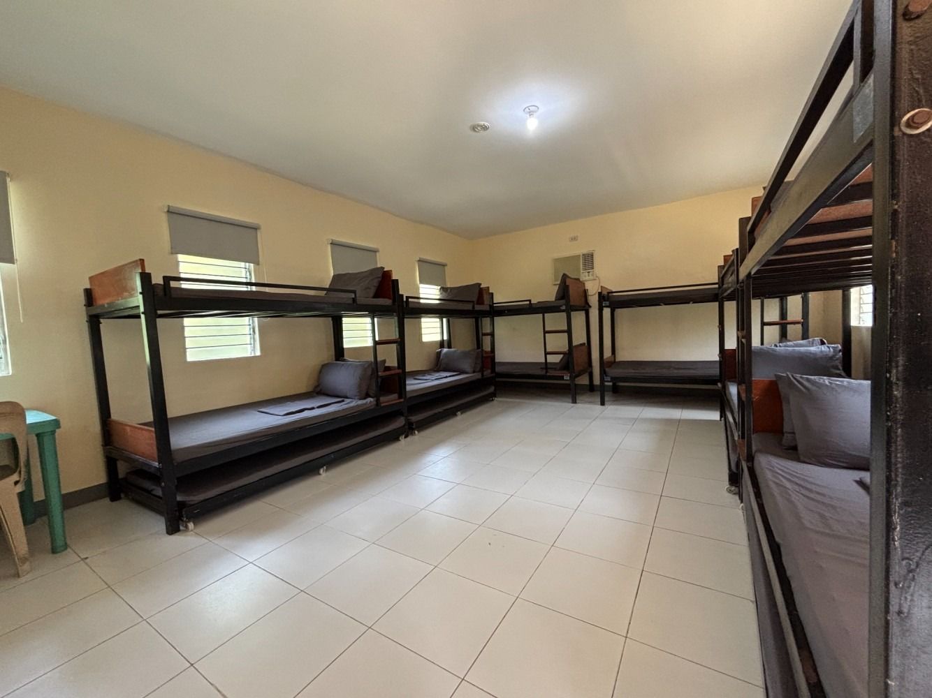 Dorm room with several bunk beds; light-colored walls and tiled floor, windows along one wall.