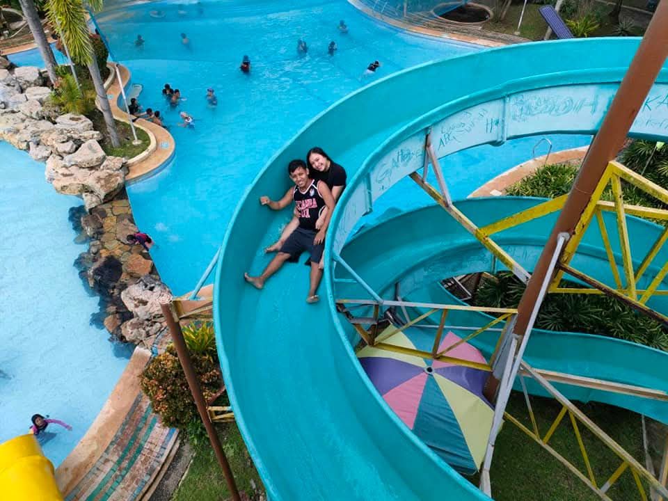Couple slides down a blue water slide into a pool. People swim nearby.