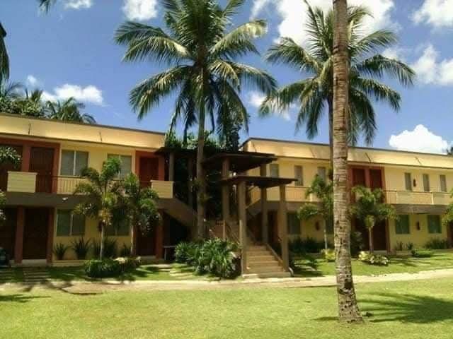 Two-story yellow building with palm trees in front, grassy lawn. Bright sunny day.