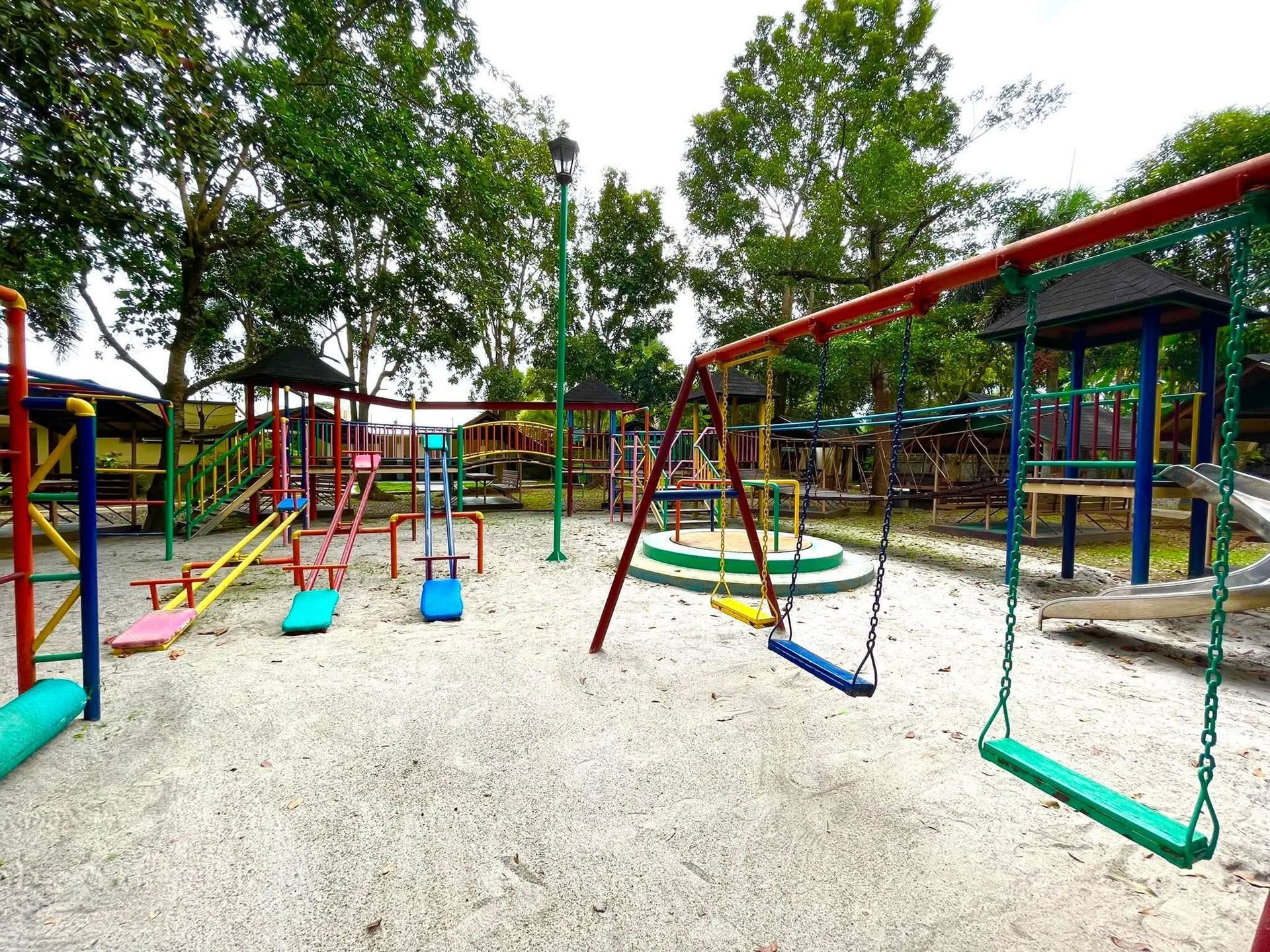 Playground with swings, seesaw, and other equipment on a gravel surface, surrounded by trees.