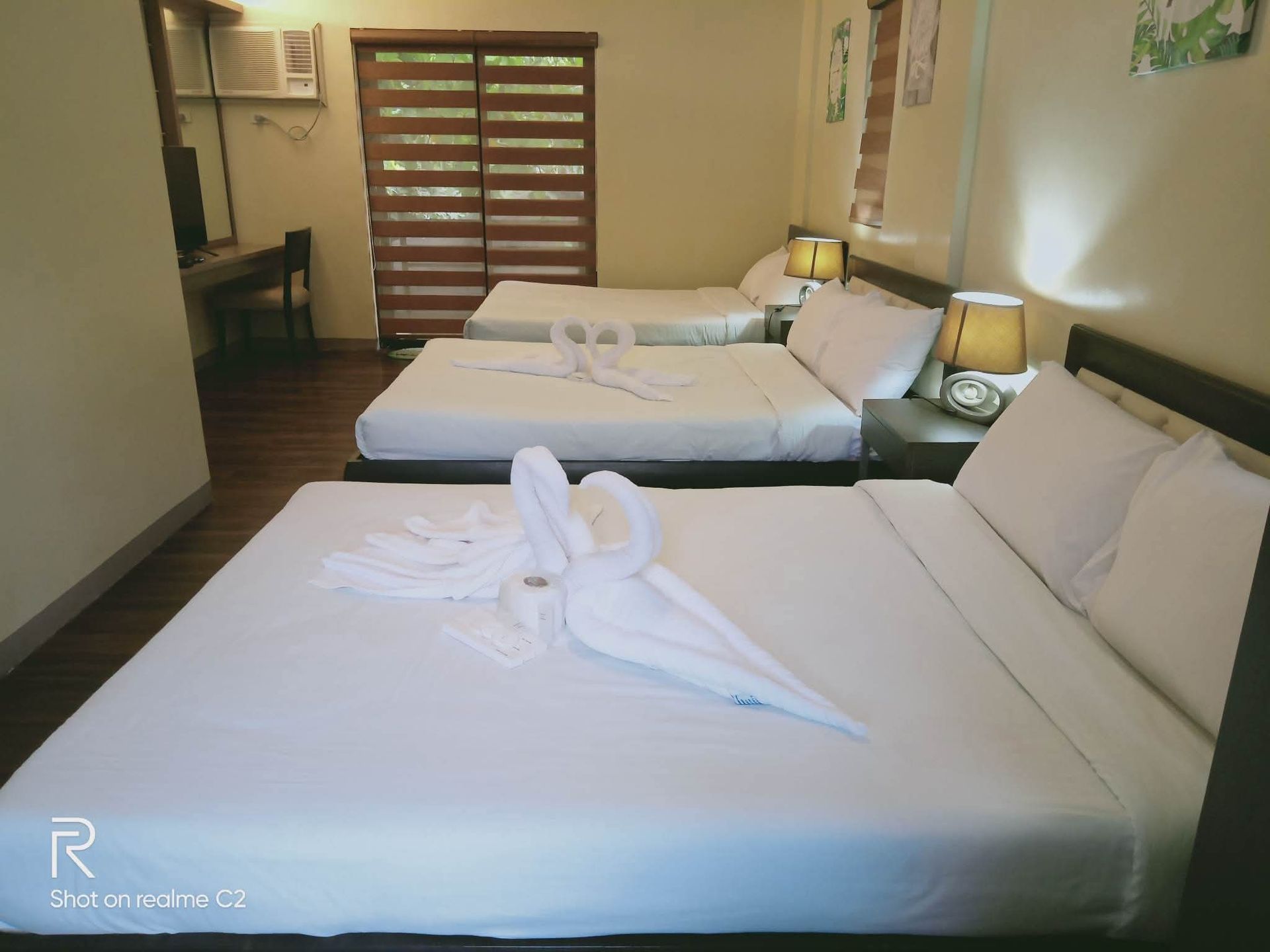 A hotel room with three beds, two with swan-shaped towel decorations, next to a window and lamps.