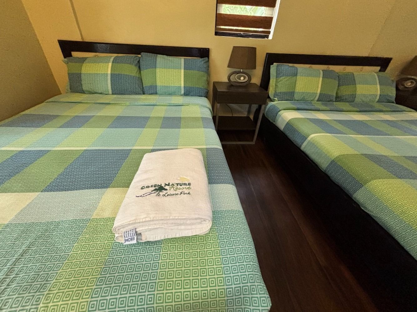 Two beds with green plaid bedding; a nightstand with a lamp; and a towel on the front bed.