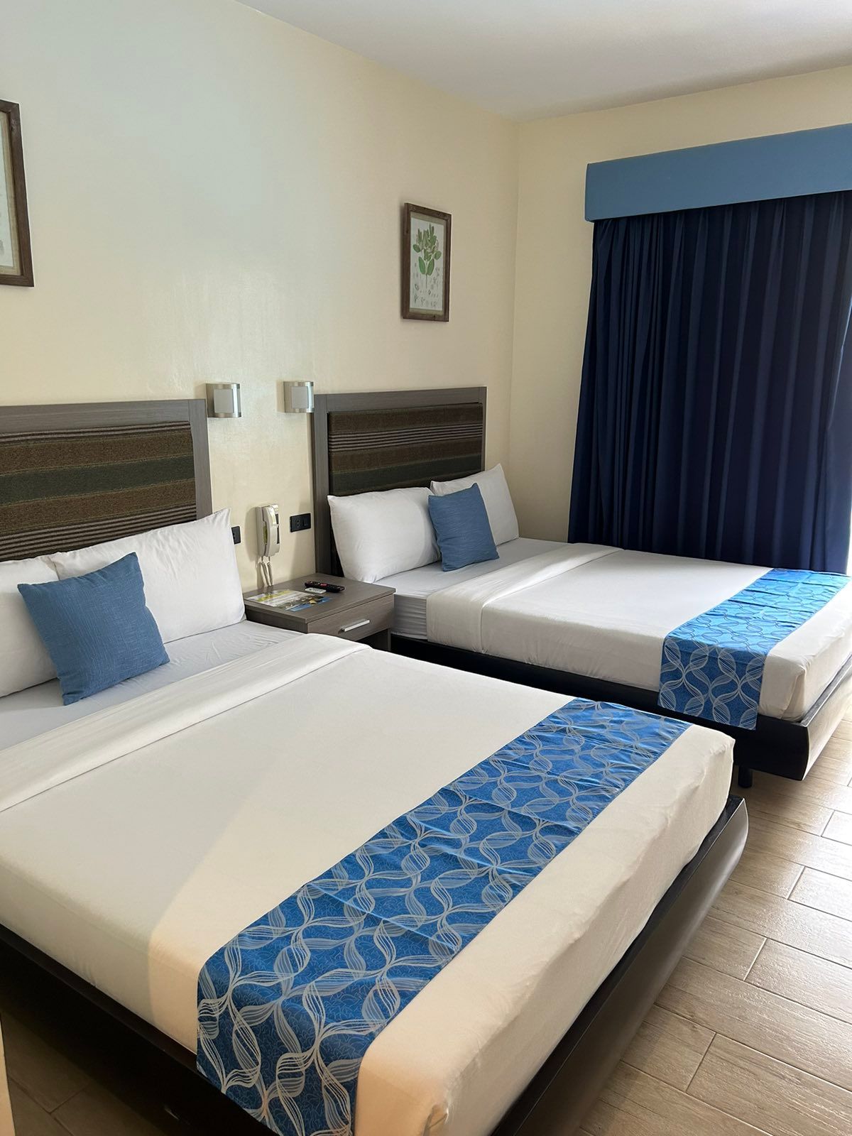 Two beds in a hotel room with blue accents; cream walls, wooden floor, and a dark blue curtain.