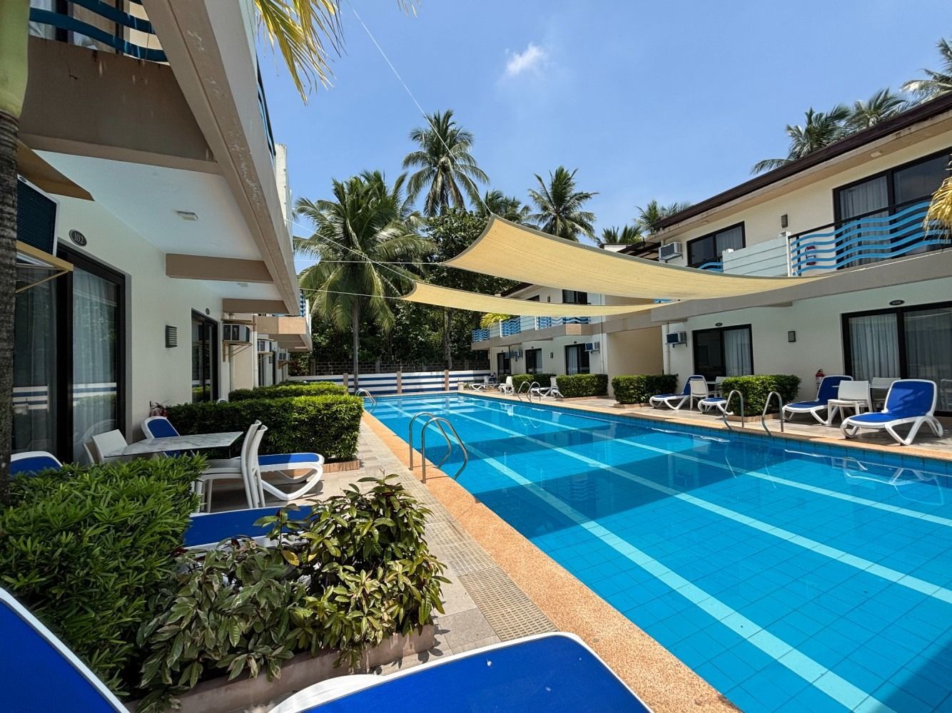 Pool with blue water and lounge chairs between two-story buildings on a sunny day.