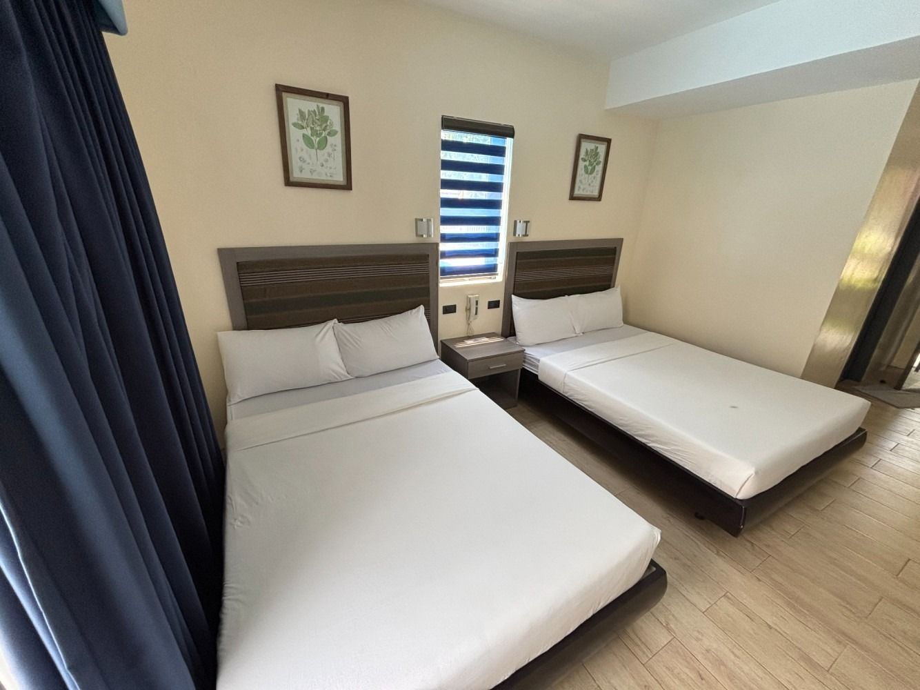 Two beds in a hotel room with white sheets, blue curtains, and framed art on the wall.