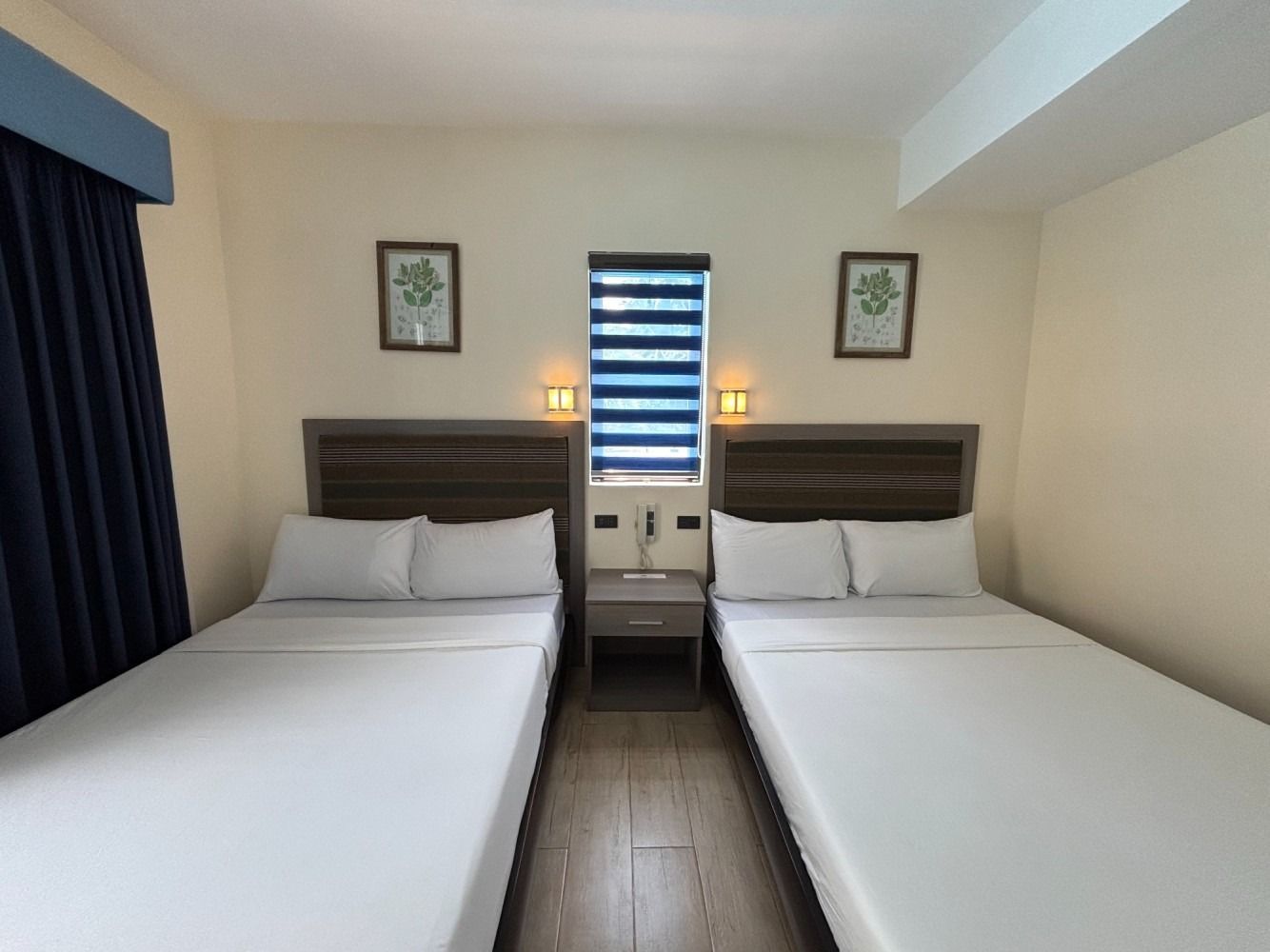 Two twin beds in a hotel room with a nightstand and framed art, cream walls, and dark blue curtains.