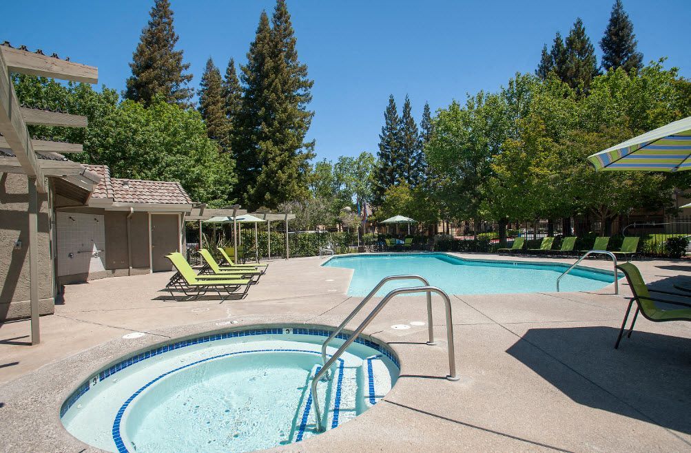 Outdoor pool and circular hot tub with green lounge chairs and trees.