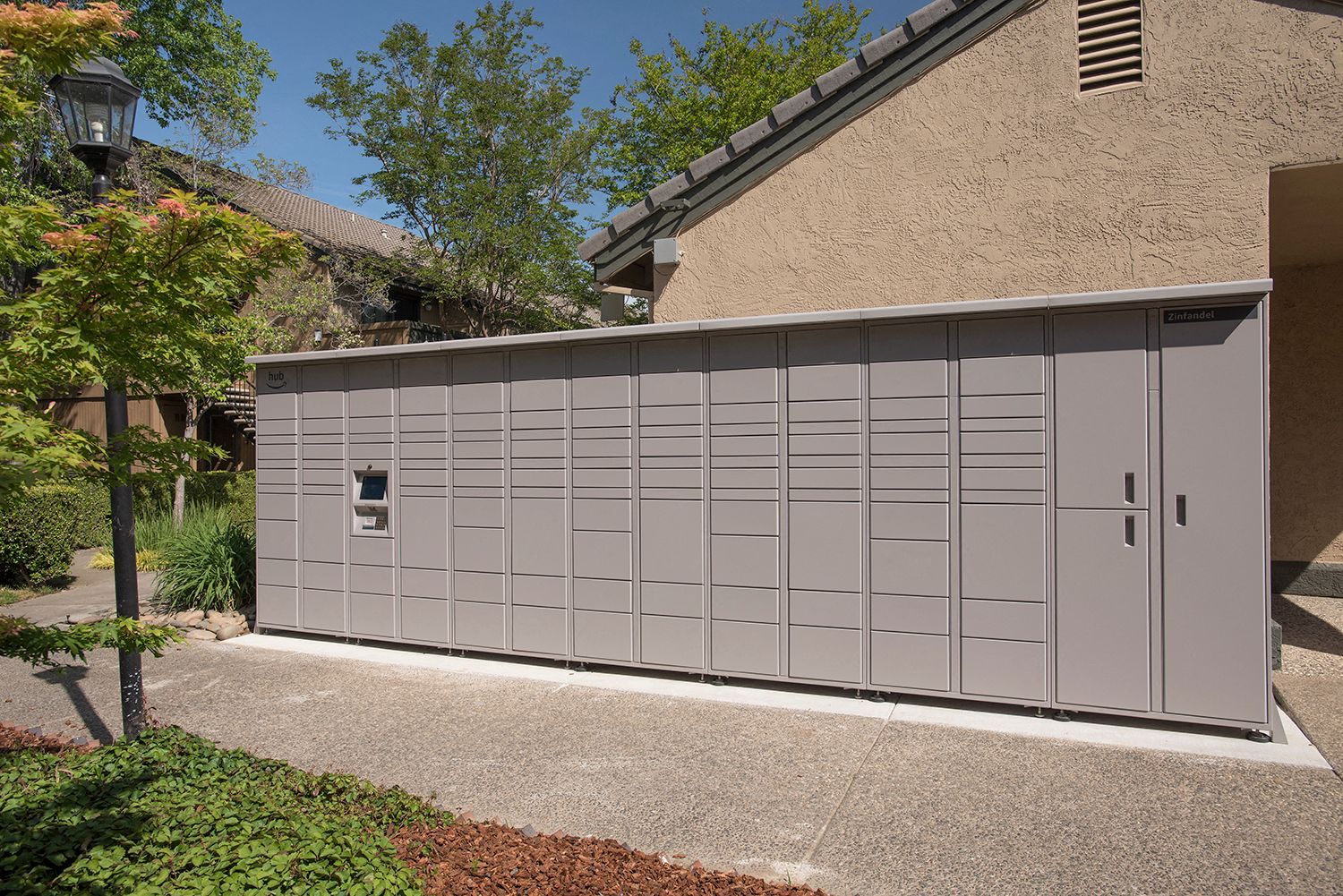 Row of gray parcel lockers along a concrete pad beside a beige stucco building.