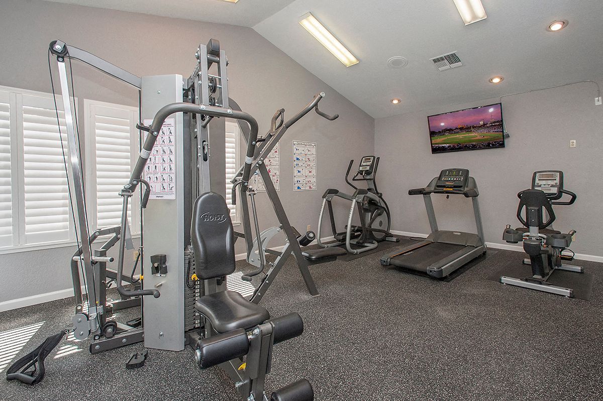 Indoor community gym with multiple cardio machines, weight station, and a wall-mounted TV.