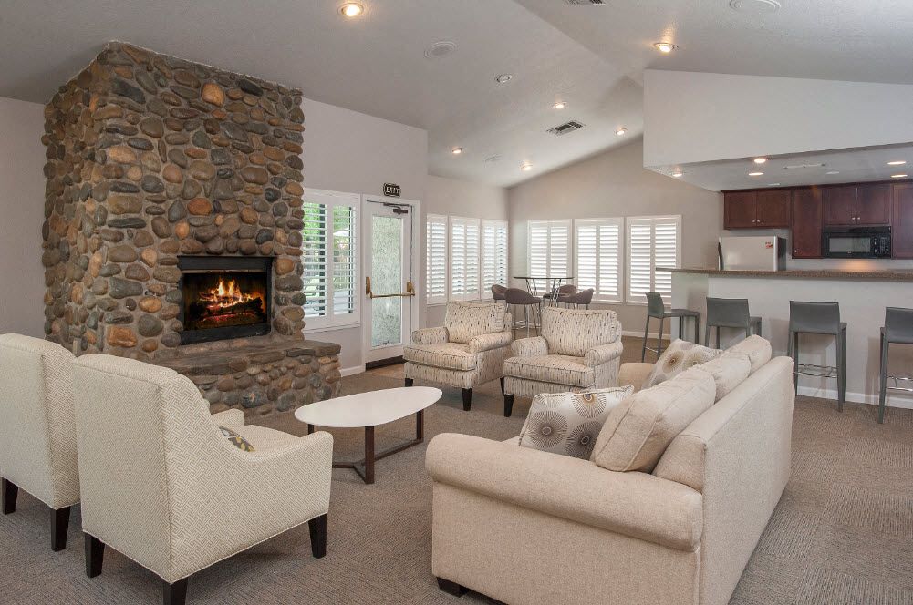 Indoor community lounge with a stone fireplace, beige seating, and a bar/kitchen area.