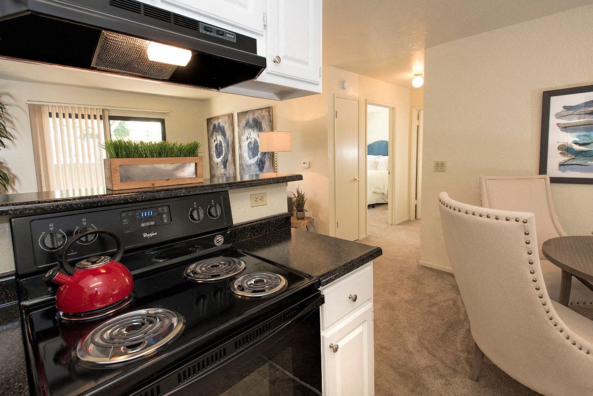 View of an apartment kitchen with a black electric stove, red kettle, white cabinets, and a dining area.
