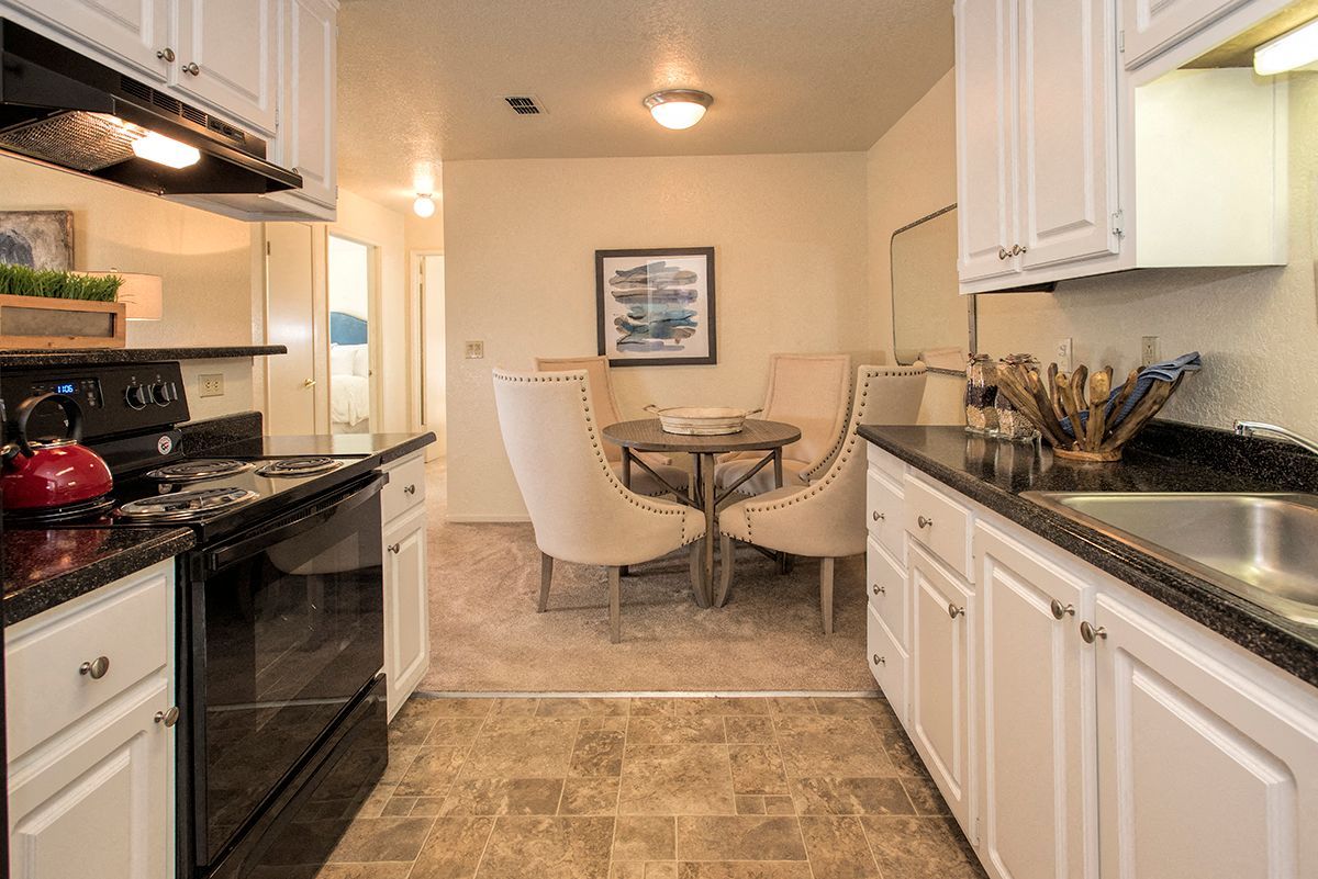 Open kitchen and dining area in an apartment, with white cabinets, black countertops, and a round table with beige upholstered chairs.