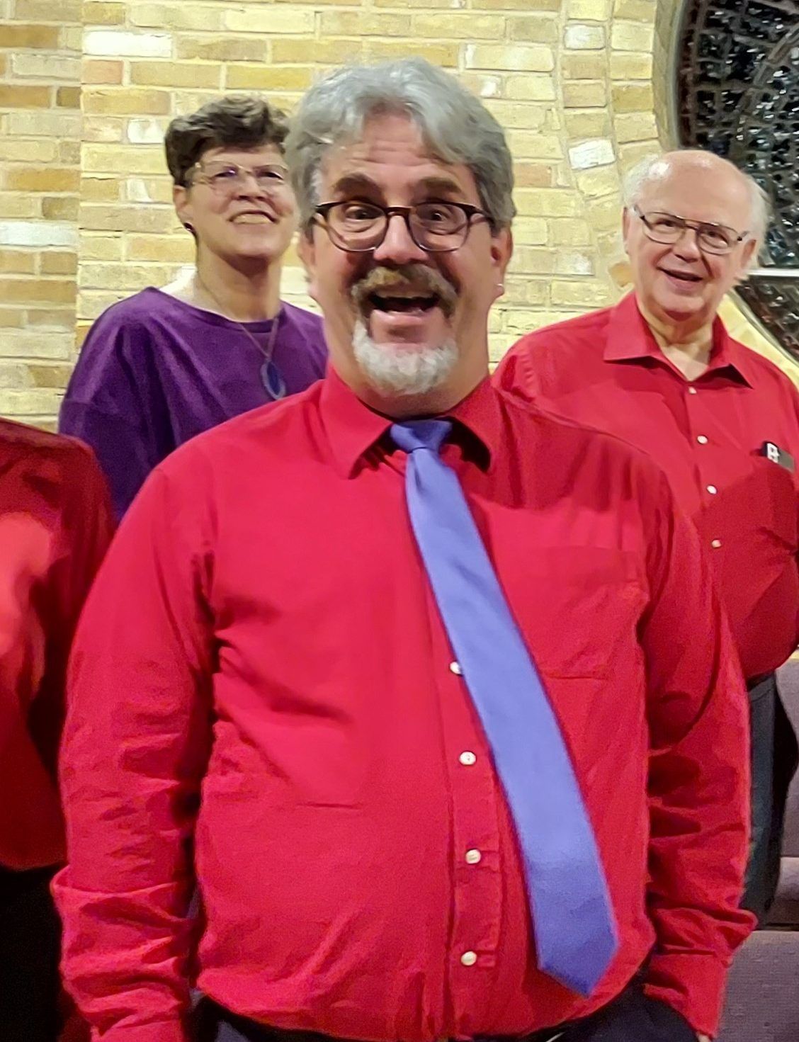 Three people in a brick-walled room; the foreground figure wears a red shirt and blue tie, smiling with an open mouth.