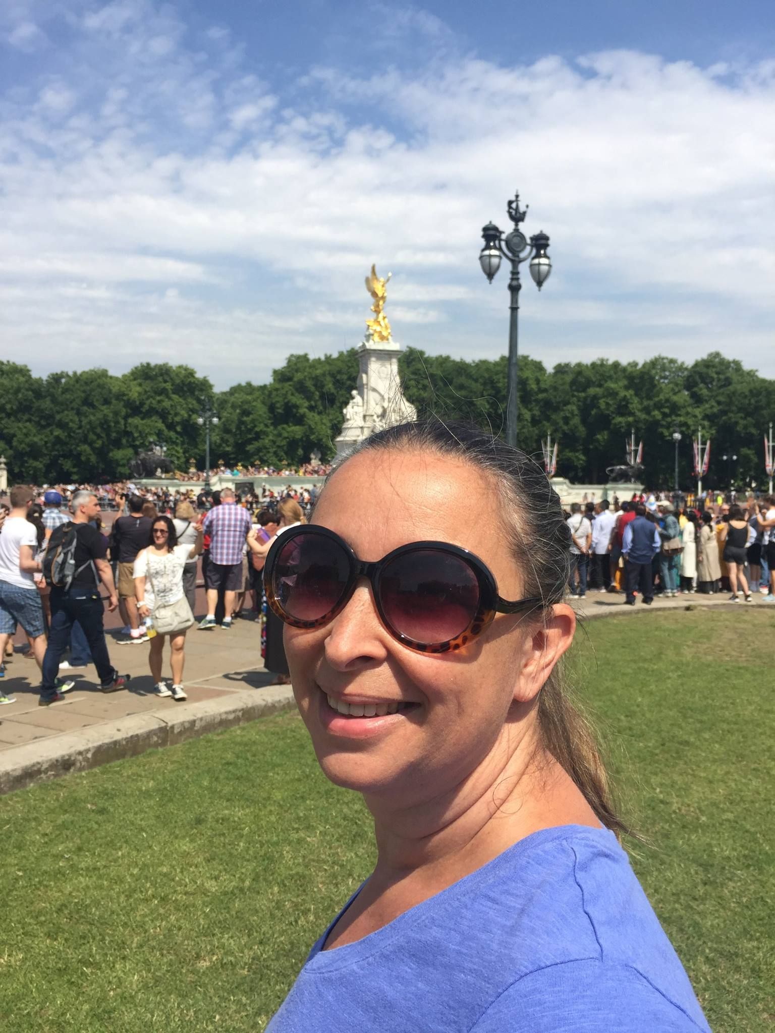 A smiling person wearing oversized sunglasses and a blue top, standing outdoors in front of the Victoria Memorial in London.